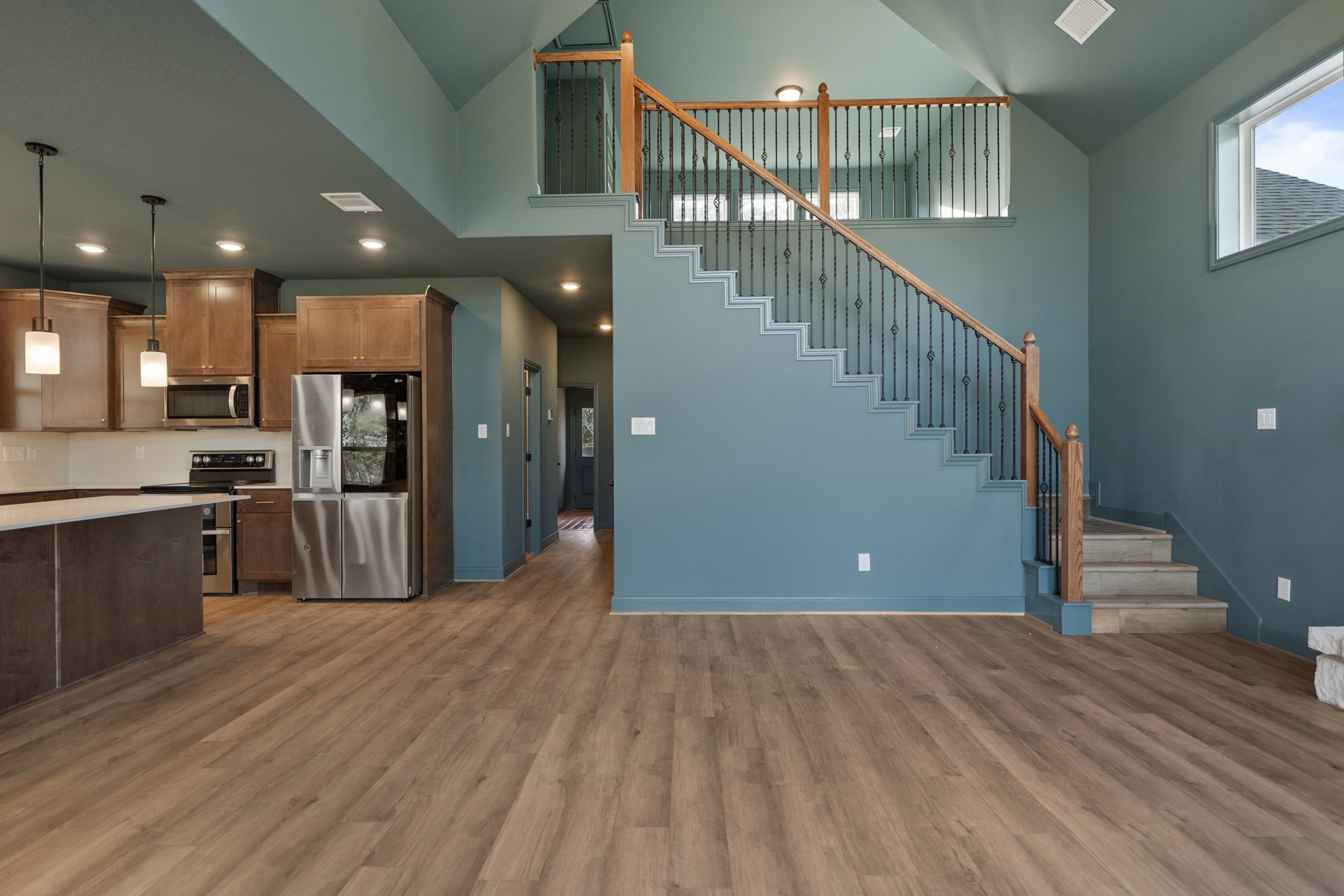 Open-concept living area featuring wood flooring, blue walls, metal-railed staircase, stainless steel refrigerator with water cooler, white vent on grey surface, and window showing