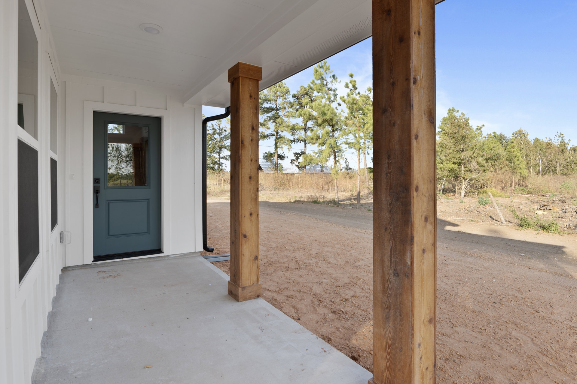 Covered porch with wooden pillars, metal roof, blue door with window, white siding, dirt road, and trees in background