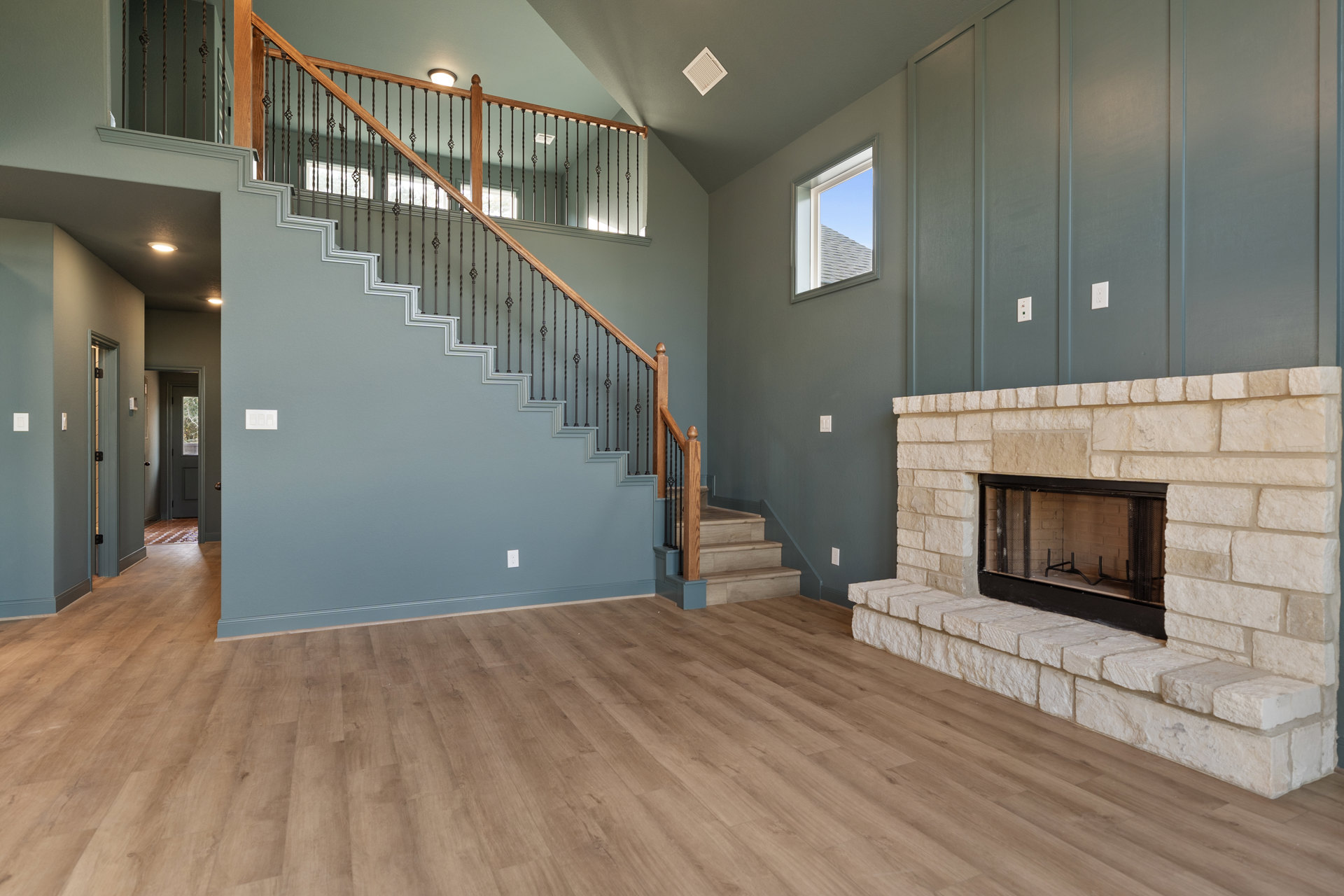 Open living room with wood flooring, modern fireplace set in a white wall, metal-railed staircase, large window showing blue sky, and grey ceiling with white vent.