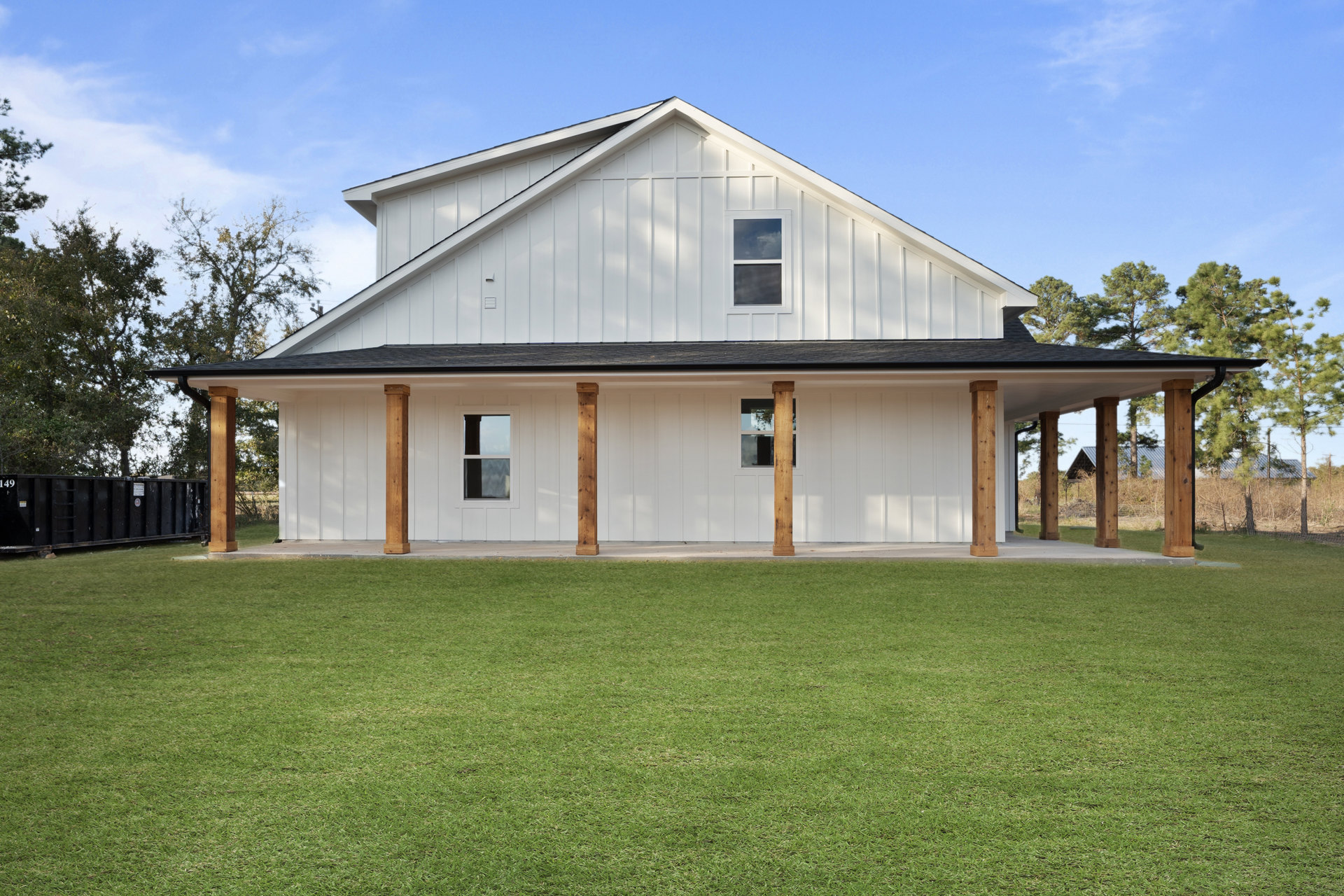 White house with wooden porch pillars, manicured lawn, blue sky reflected in front window, American Gothic House visible in background, black dumpster near property.