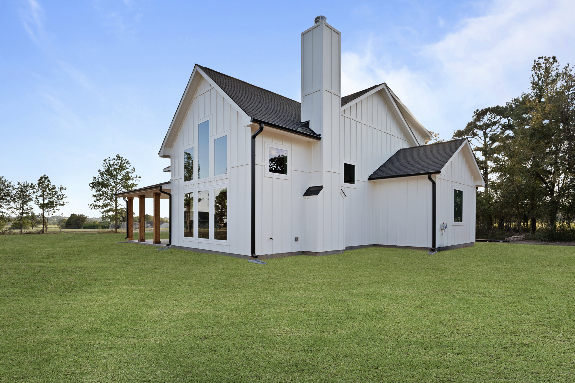 White farmhouse with black chimney, rain gutters, and large windows, surrounded by green lawn and mature tree under partly cloudy sky