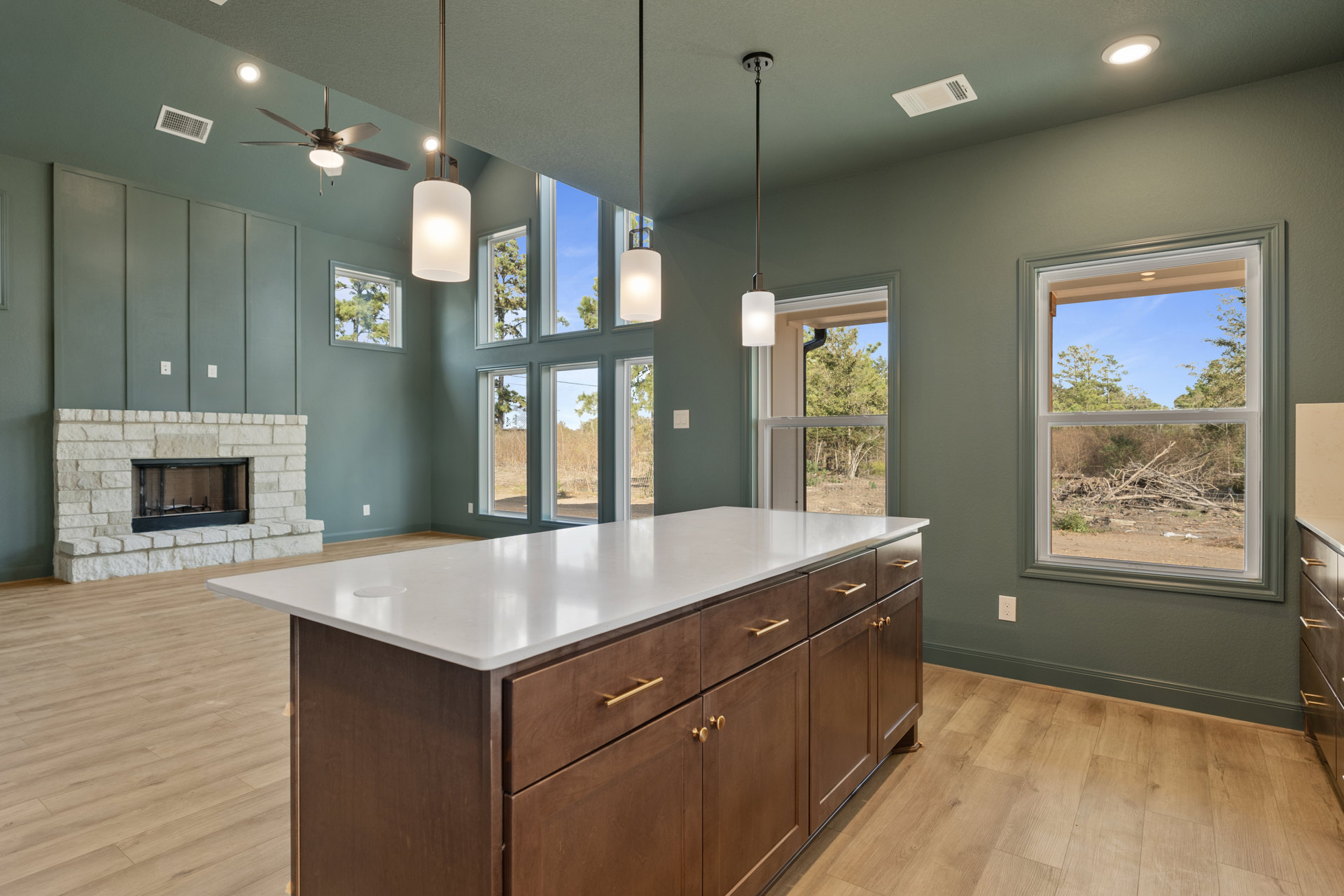 Kitchen island with white countertop and built-in drawers, light fixture overhead, cabinetry and sink, large window overlooking forested landscape, fireplace with glass door on