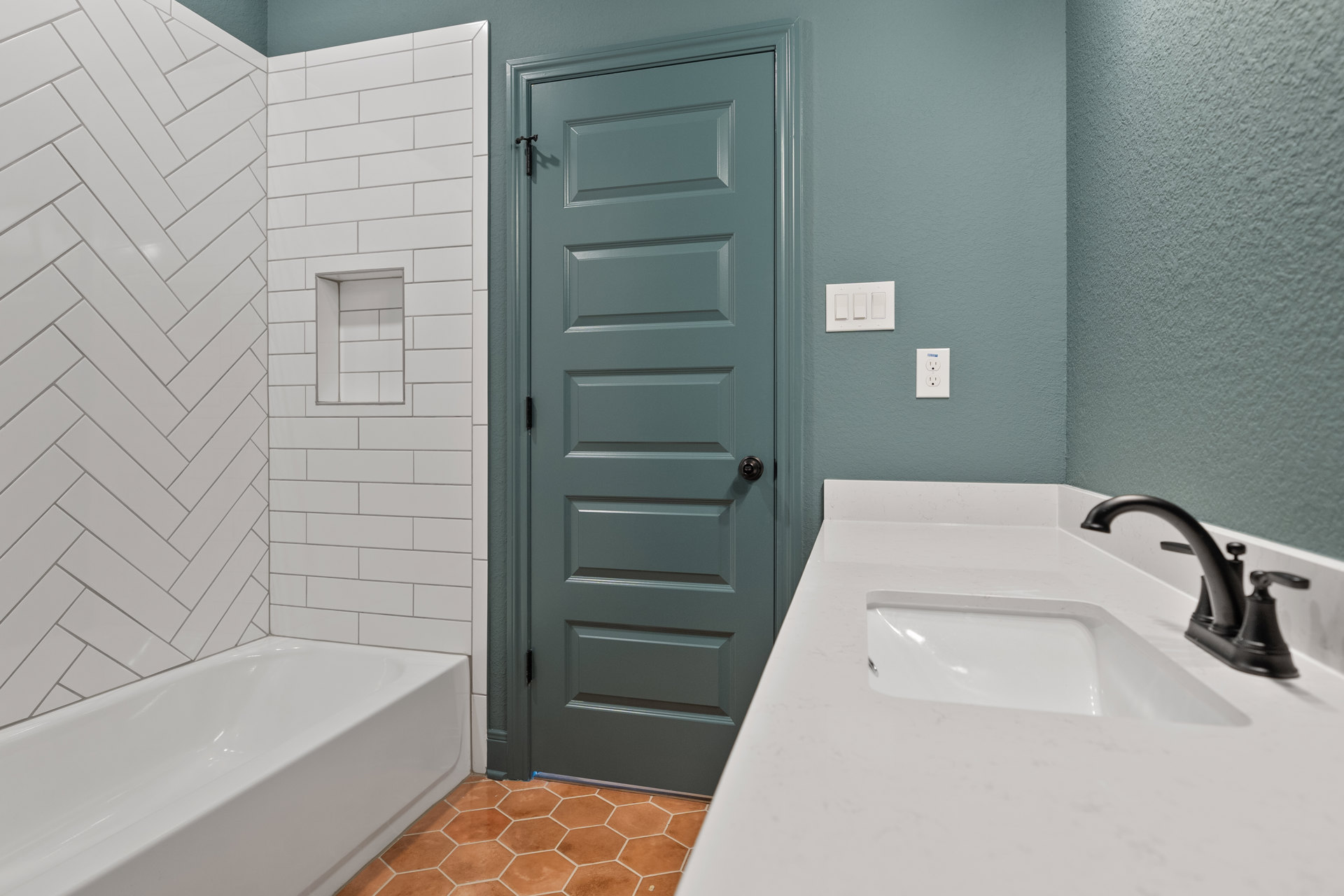White bathroom with rectangular sink and chrome faucet, freestanding white bathtub against tiled wall with built-in shelf, white door with silver knob, and white light switch on