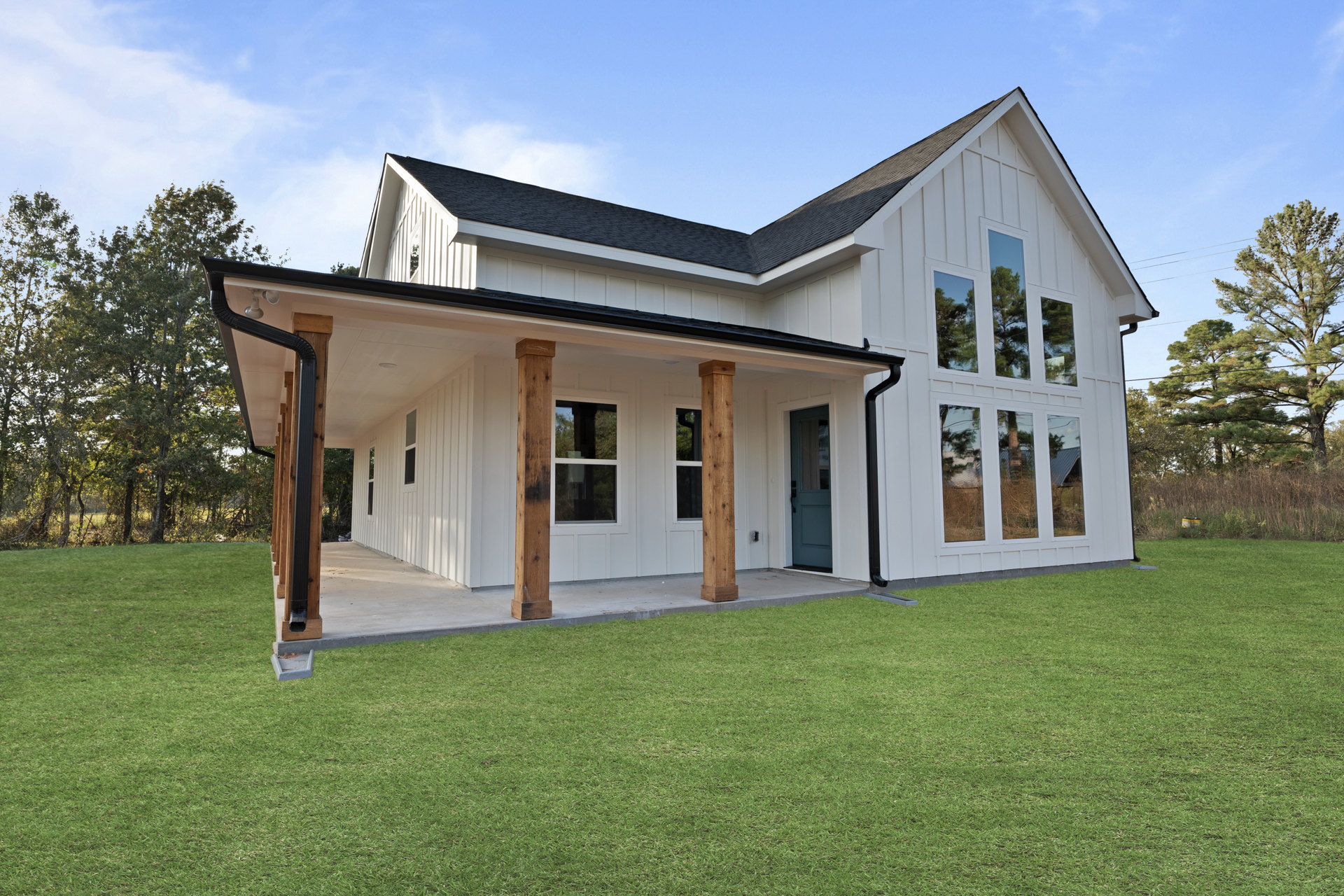 White house with blue door, wooden porch pillars, large windows reflecting trees, surrounded by green grass lawn and landscaping.