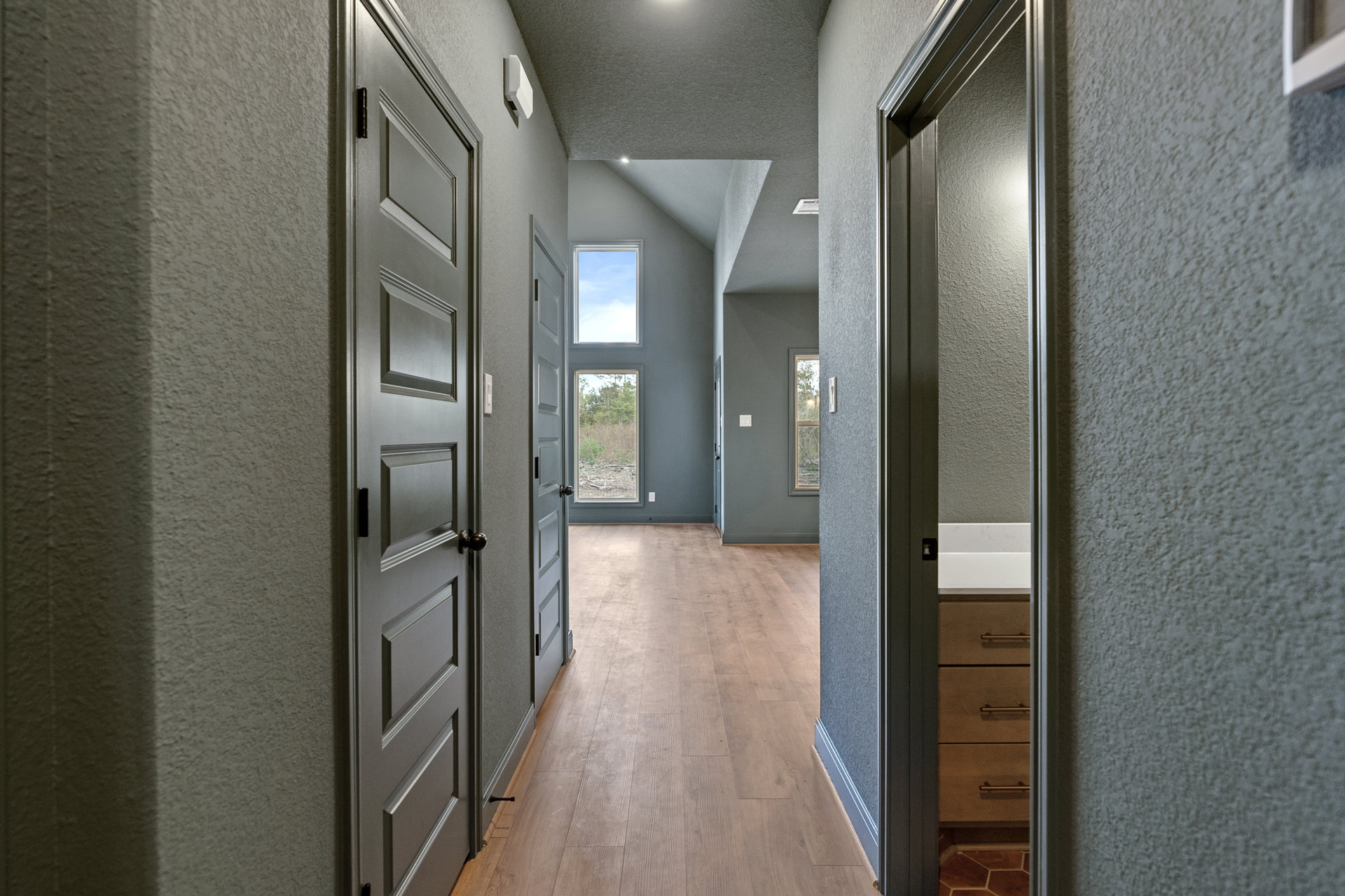 Hallway with smooth grey walls, light wood flooring, white doors, and a window showing blue sky and green trees; cabinetry with drawers visible along one side