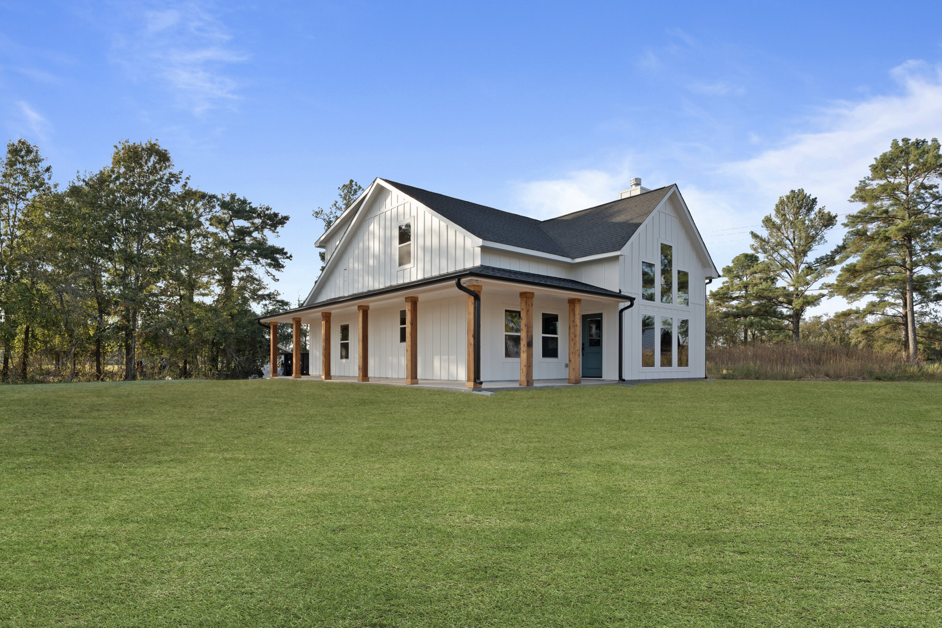 White house with black roof and wood pillars, surrounded by green lawn and tall trees under partly cloudy sky