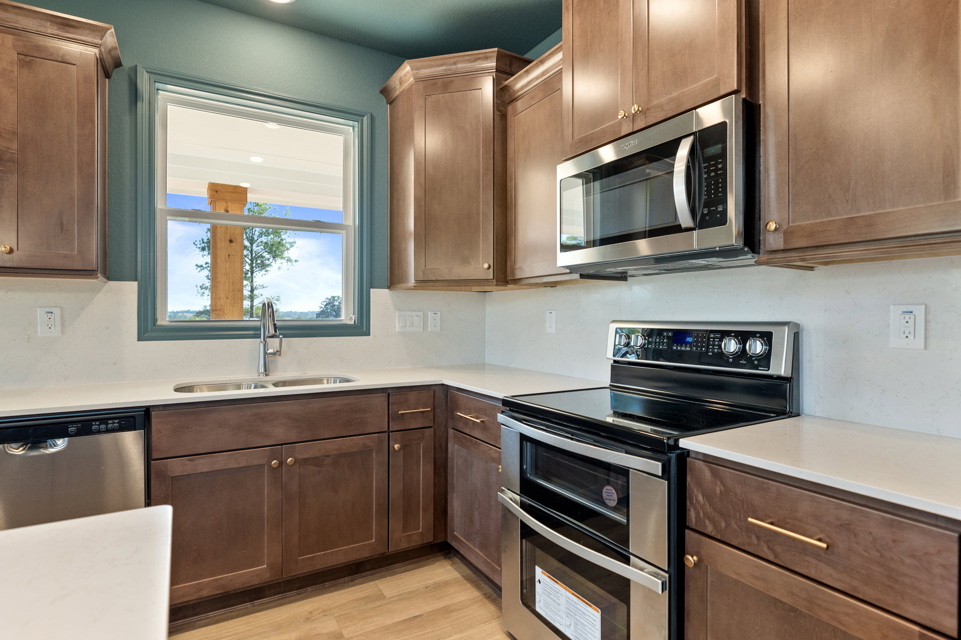 Modern kitchen featuring stainless steel oven with glass door, built-in microwave in wood cabinetry, white countertops, sink beneath window overlooking tree, and stove with metal
