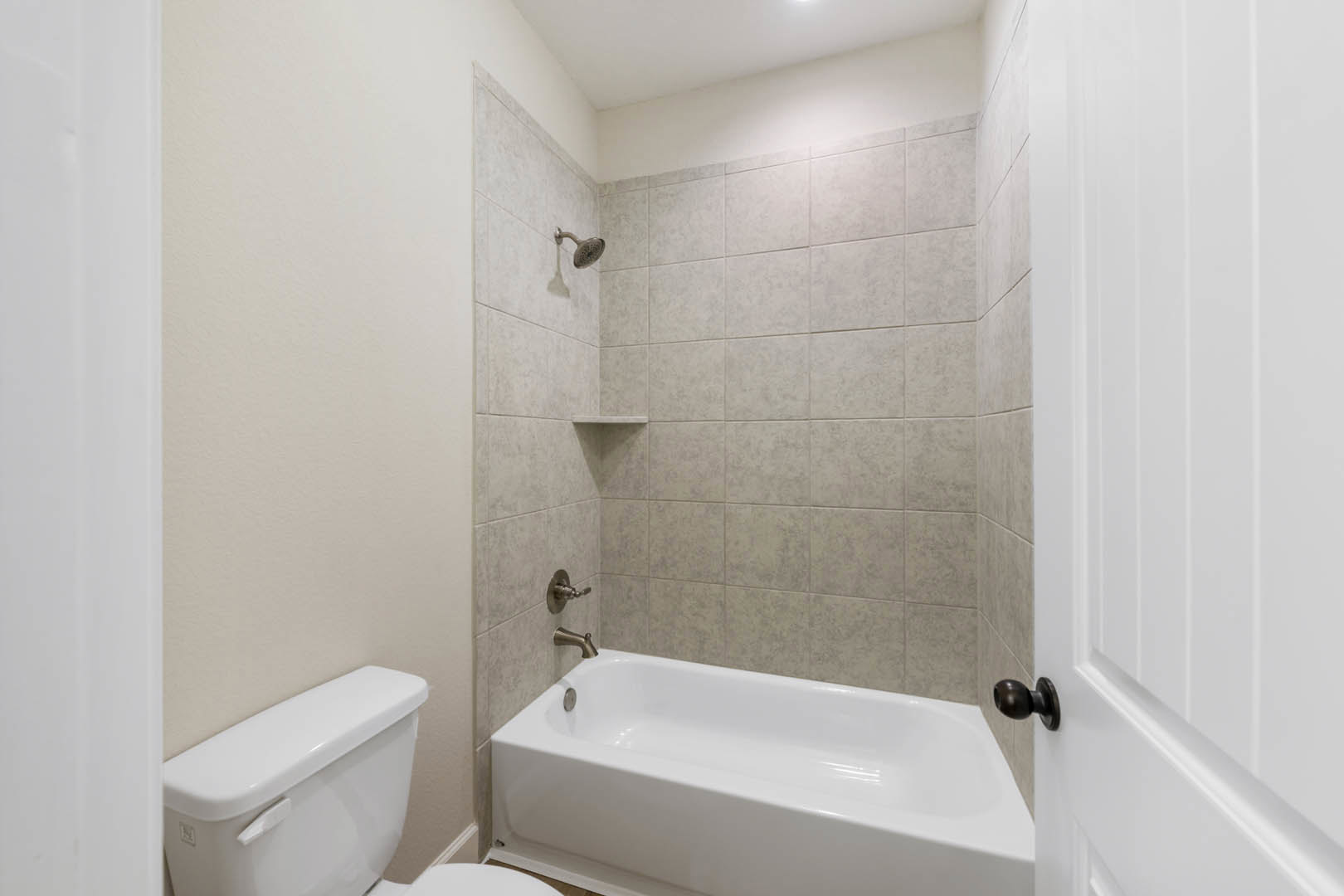 White bathtub set against a brown tiled accent wall, adjacent to a white toilet; chrome faucet and handle visible, with white walls and tile flooring.