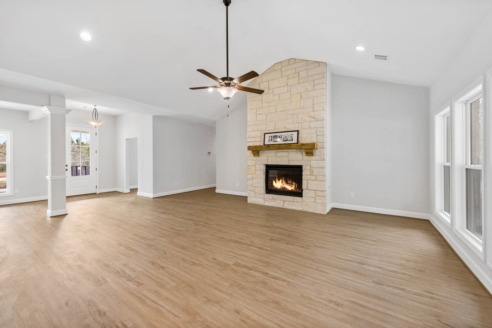 Living room with hardwood floors, stone fireplace with burning fire, ceiling fan with lights, neutral walls, and large windows letting in natural light