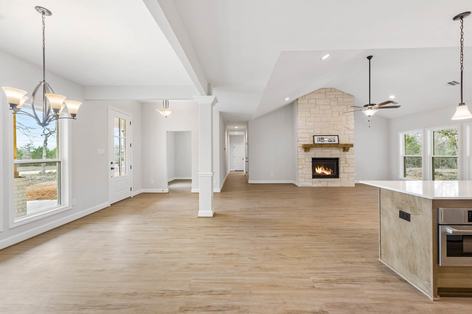 Spacious living room featuring wood flooring, white pillars, active fireplace, ceiling fan, and large windows beneath a white ceiling with decorative columns