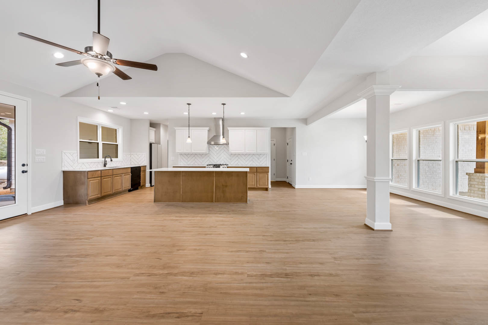 Spacious open-concept interior featuring a central kitchen island with white cabinetry, wood flooring, ceiling fan with light fixture, white walls, open doorway, and large window