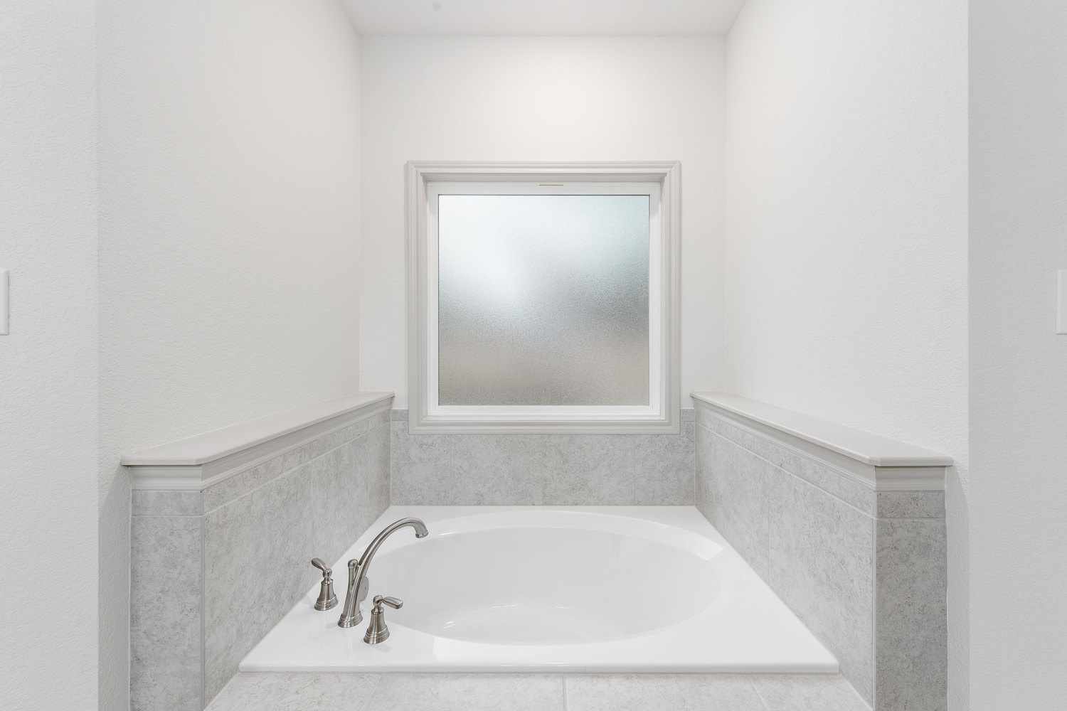 White freestanding bathtub with chrome faucet set against frosted glass window, surrounded by light tile flooring and white walls in a modern bathroom.