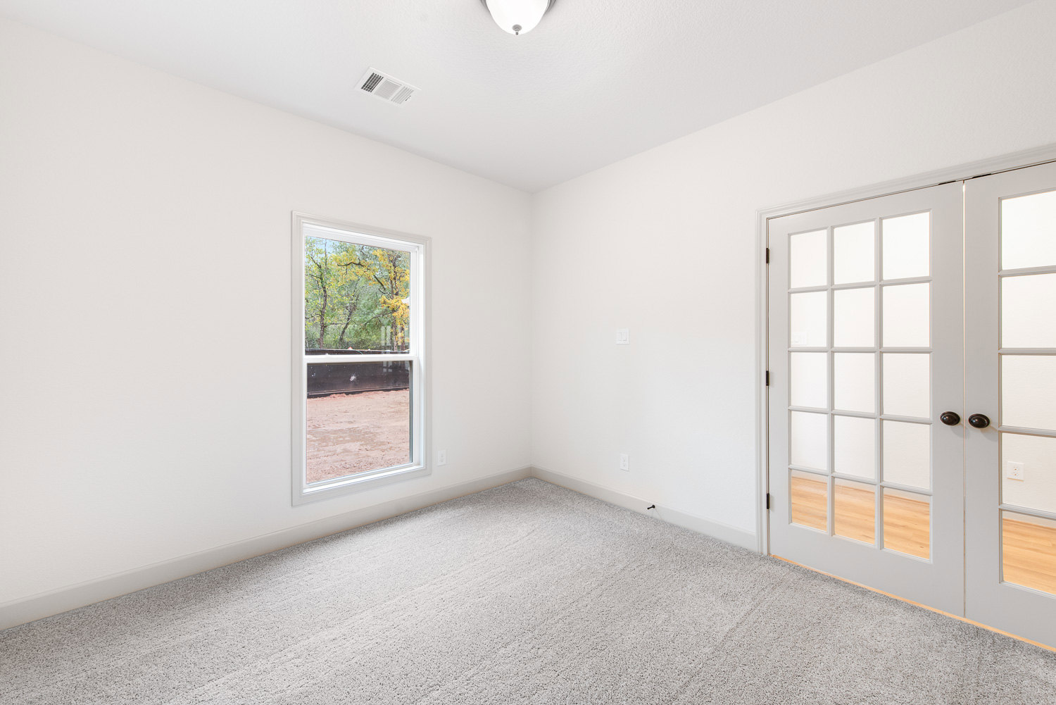 White carpeted room featuring a white door with black handles, large window overlooking trees and dirt, white vent on wall.