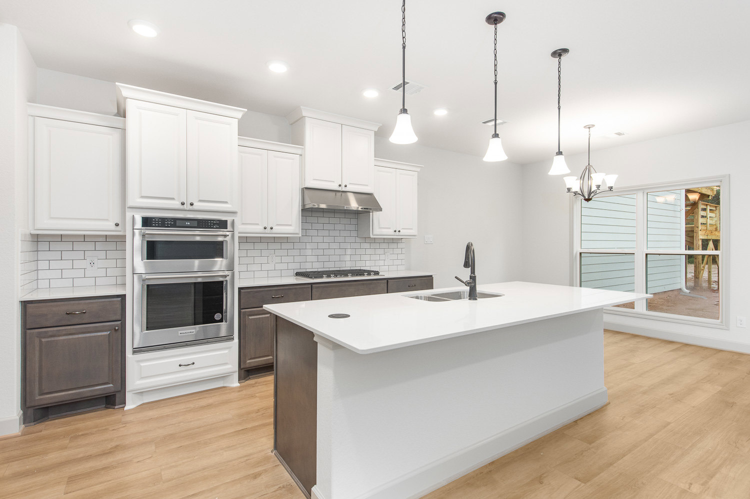 White kitchen with shaker-style cabinets, white island featuring built-in sink, stainless steel oven, hardwood flooring, and pendant light with chain above island.