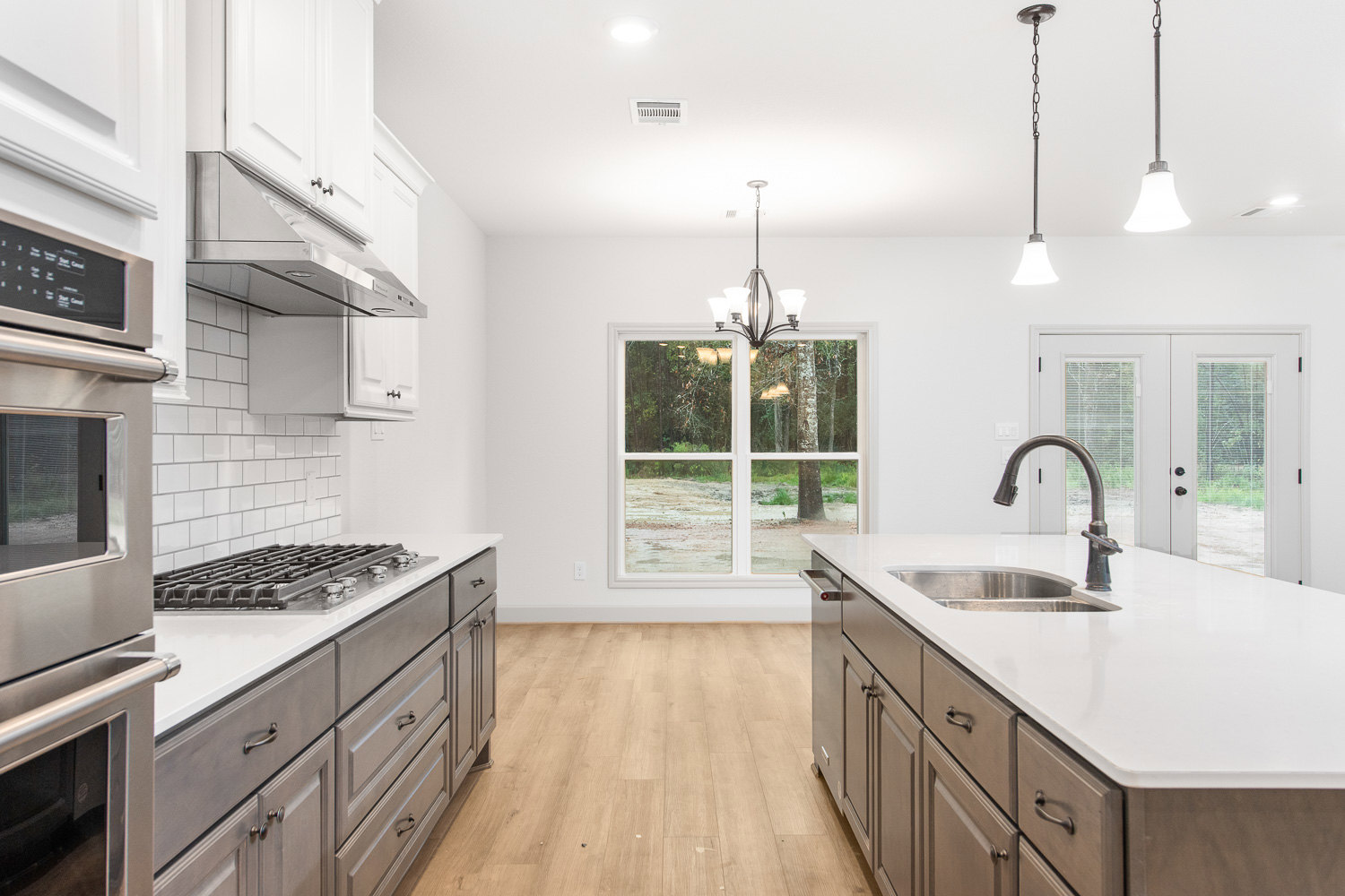 Spacious kitchen featuring a large central island with built-in sink, white tile backsplash beneath a window overlooking a forest, modern cabinetry, pendant lighting, and stainless