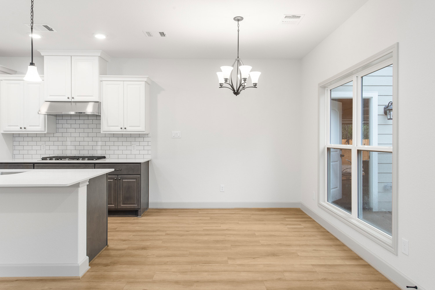 Kitchen with light wood flooring, white cabinetry with silver knobs, white countertops, stainless steel sink, modern chandelier, and large window framed in white.