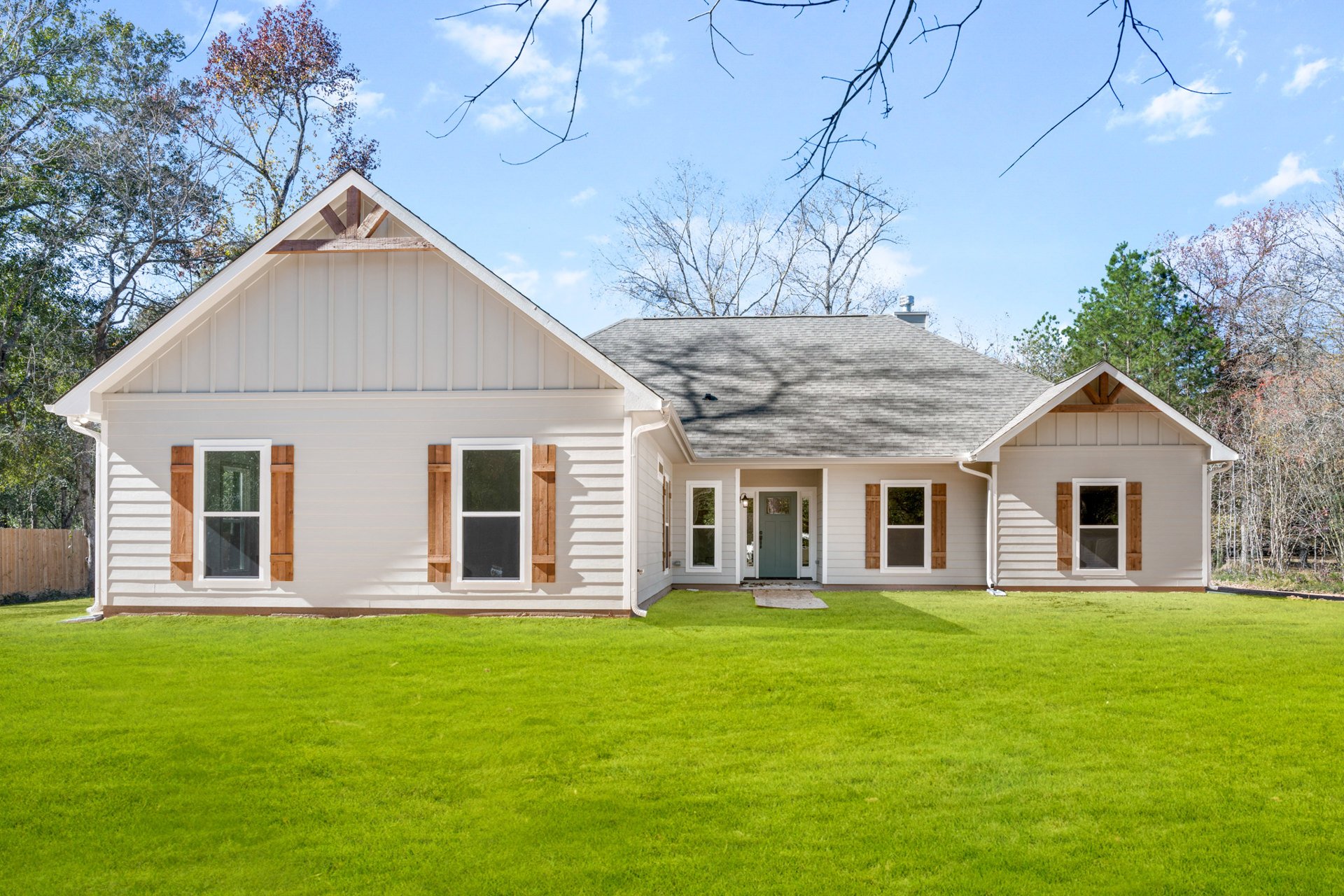 Two-story house with white-framed windows, green front door with glass panel, gray shingle roof, manicured lawn, mature trees in background, blue sky with scattered clouds