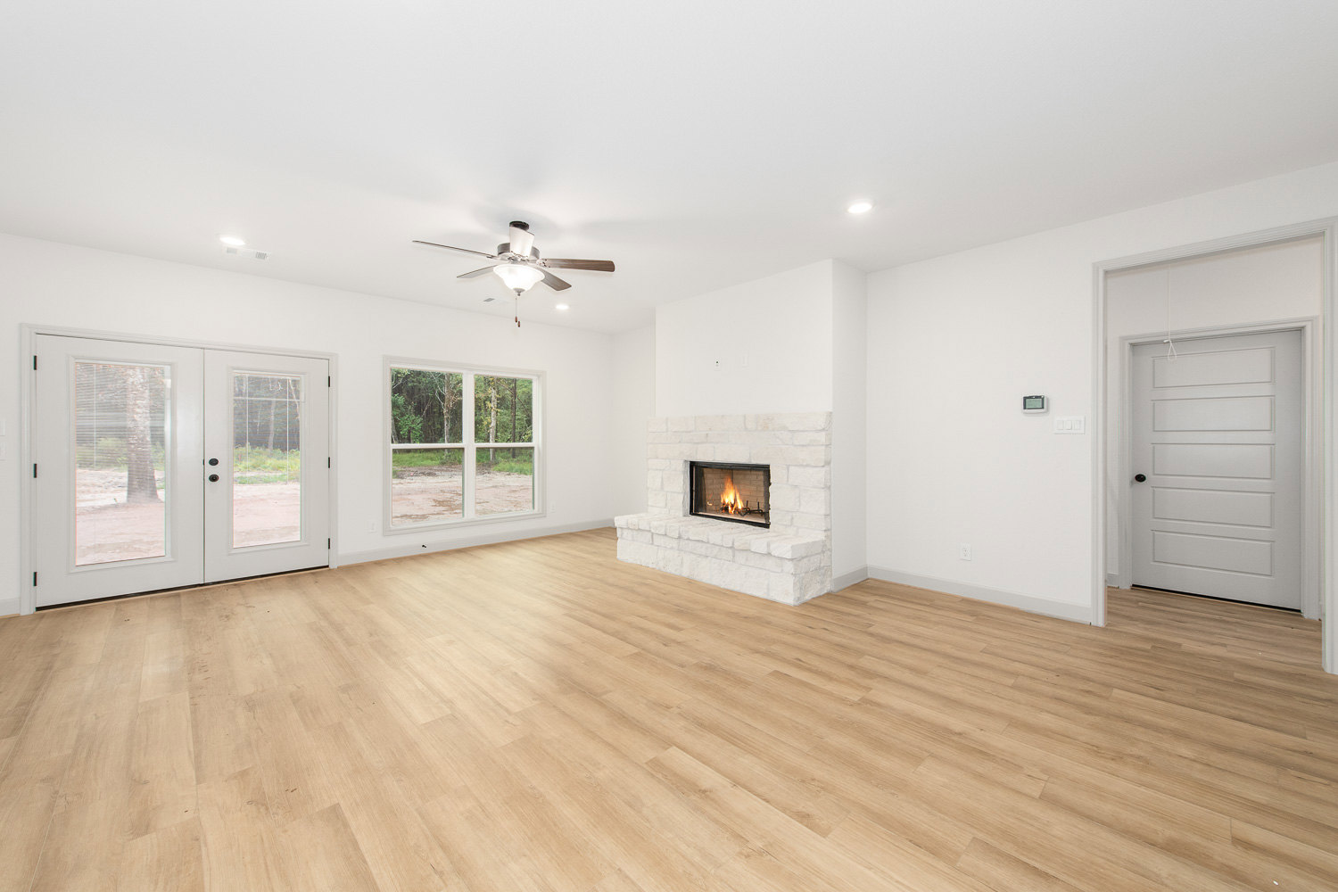 Living room with hardwood floors, active fireplace with stone surround, white door with black knob, ceiling fan with light fixture, plaster walls.