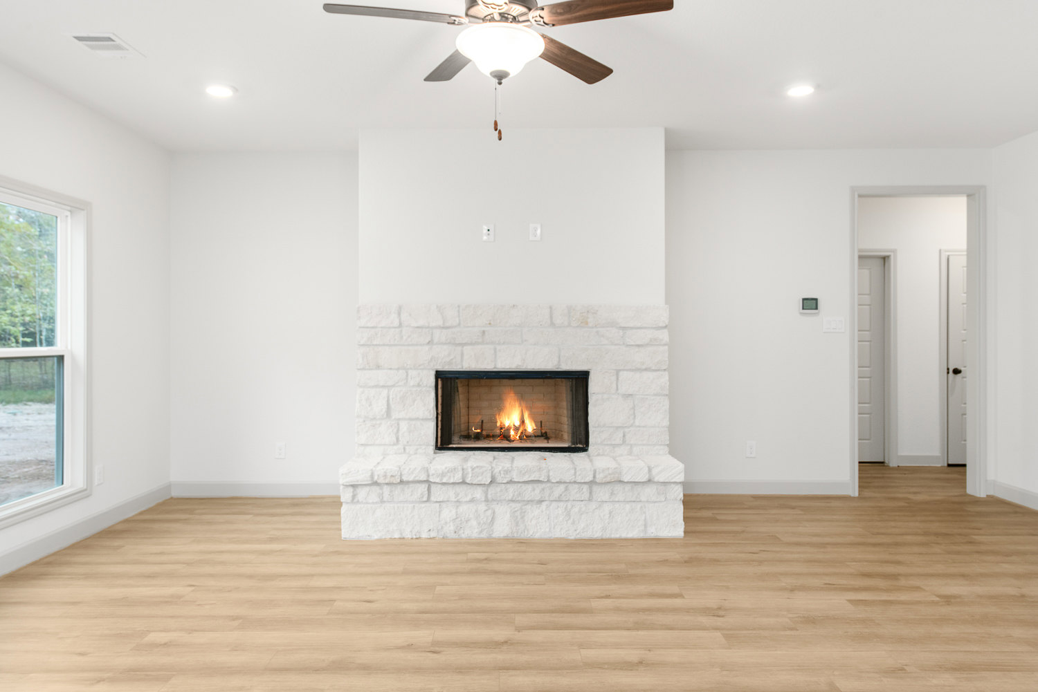 Fireplace with burning fire set in a white brick wall, wood flooring, ceiling fan with light fixture overhead, window revealing trees outside
