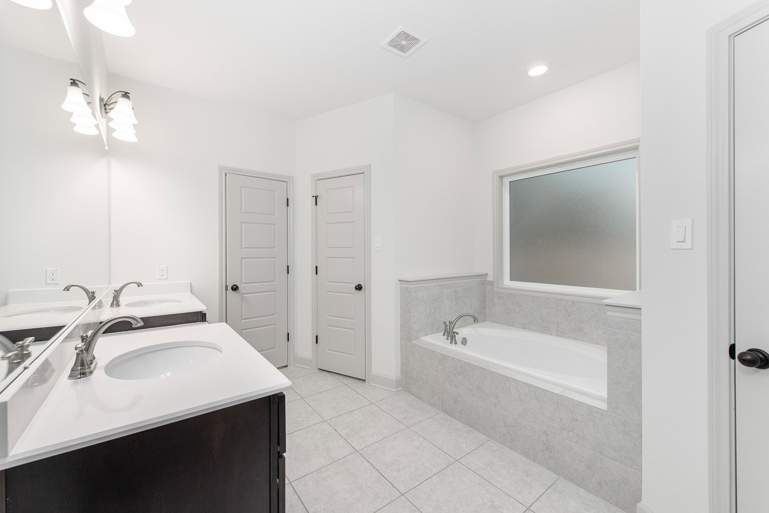 Freestanding bathtub beside white tile wall, modern sink with chrome faucet, white door with black hardware, window with trim, and contemporary light fixture
