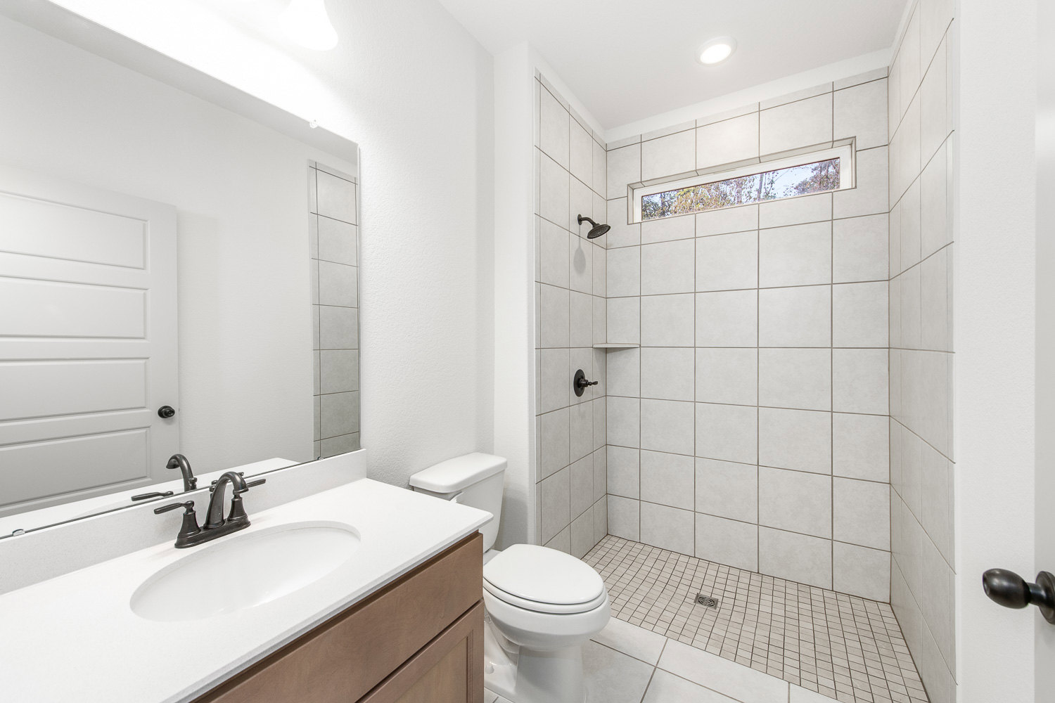 Modern bathroom featuring a glass-enclosed shower, white toilet with lid down beside a stone countertop and undermount sink, rectangular window, ceiling light, and neutral tile