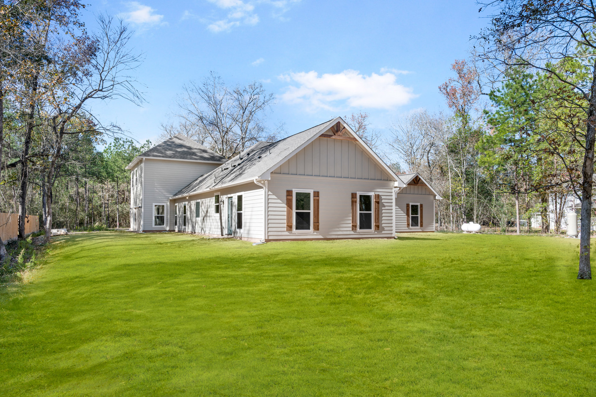 Two-story house with gray siding, white trim windows, shingled roof, and manicured green lawn bordered by mature trees under a partly cloudy sky