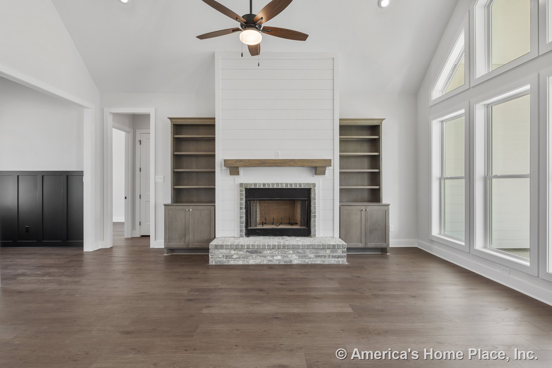Central brick fireplace with wood mantel set against a shiplap accent wall, flanked by built-in shelves and cabinets; vaulted ceiling with ceiling fan, wide plank wood flooring