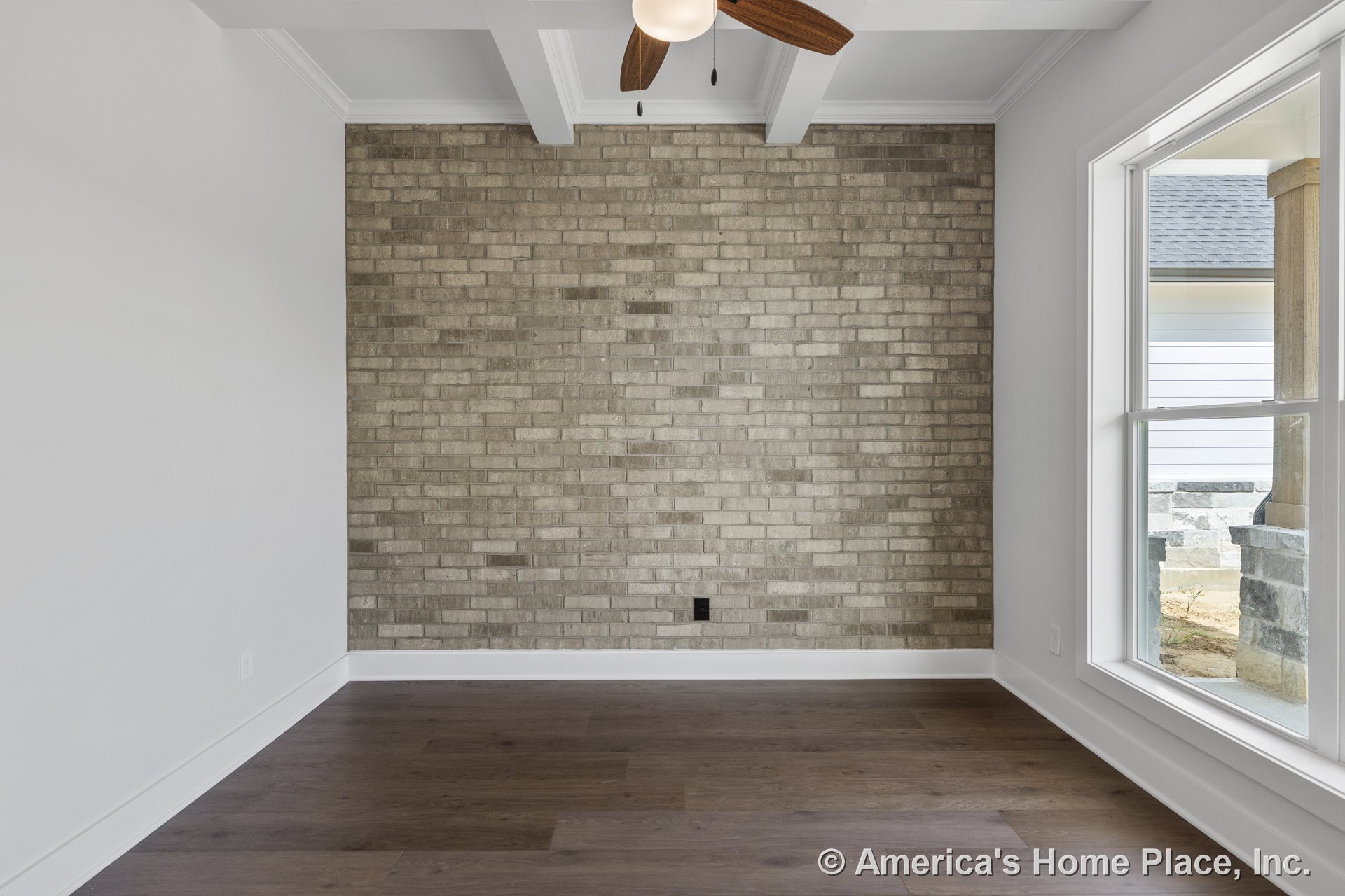 Brick accent wall, large window with white trim, wood plank flooring, ceiling fan with light, exposed ceiling beams, and white baseboards in a modern, naturally lit room with