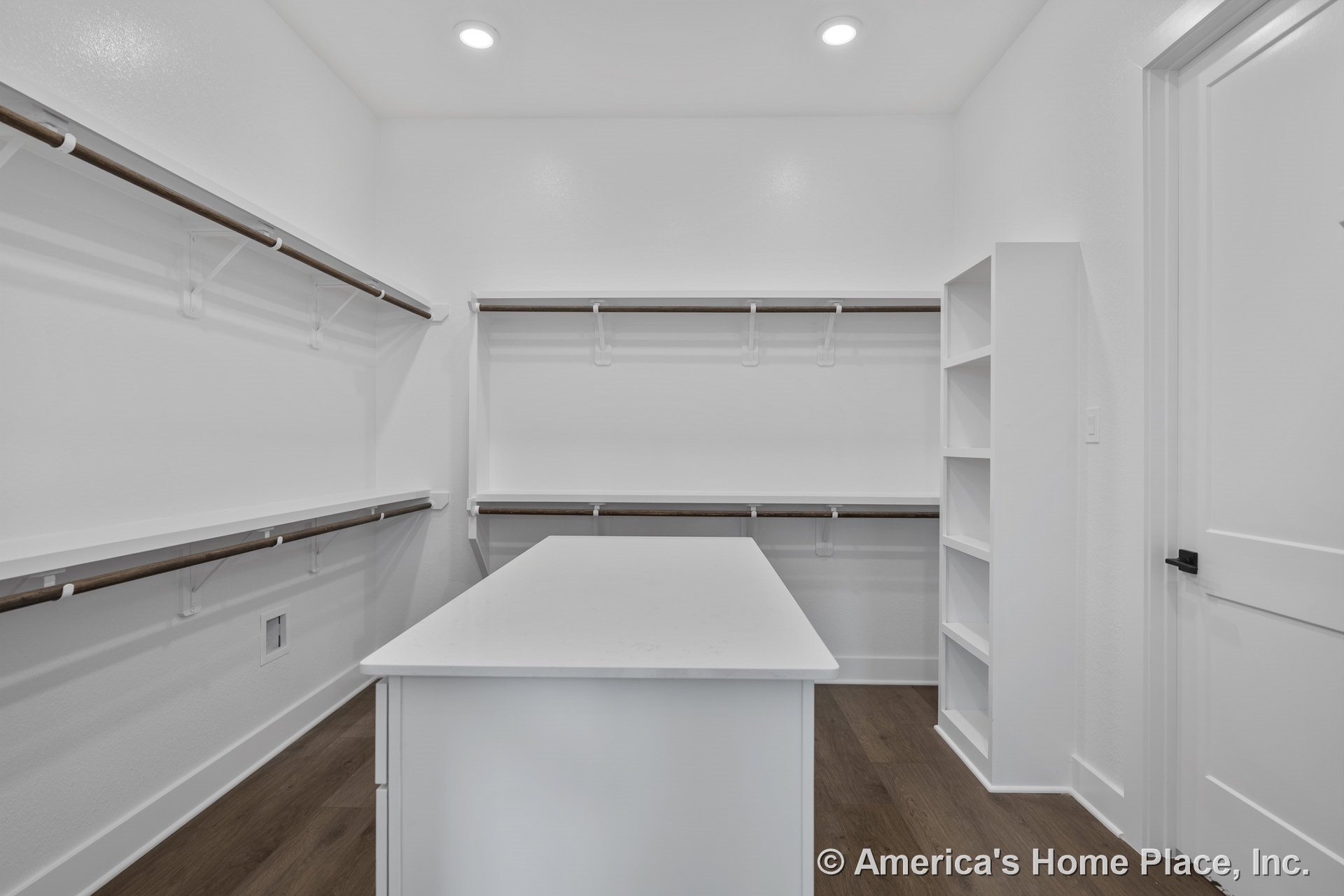 Walk-in closet with built-in shelving, wood hanging rods, central island topped with white countertop, recessed ceiling lights, wood-look flooring, white trim, and interior door.