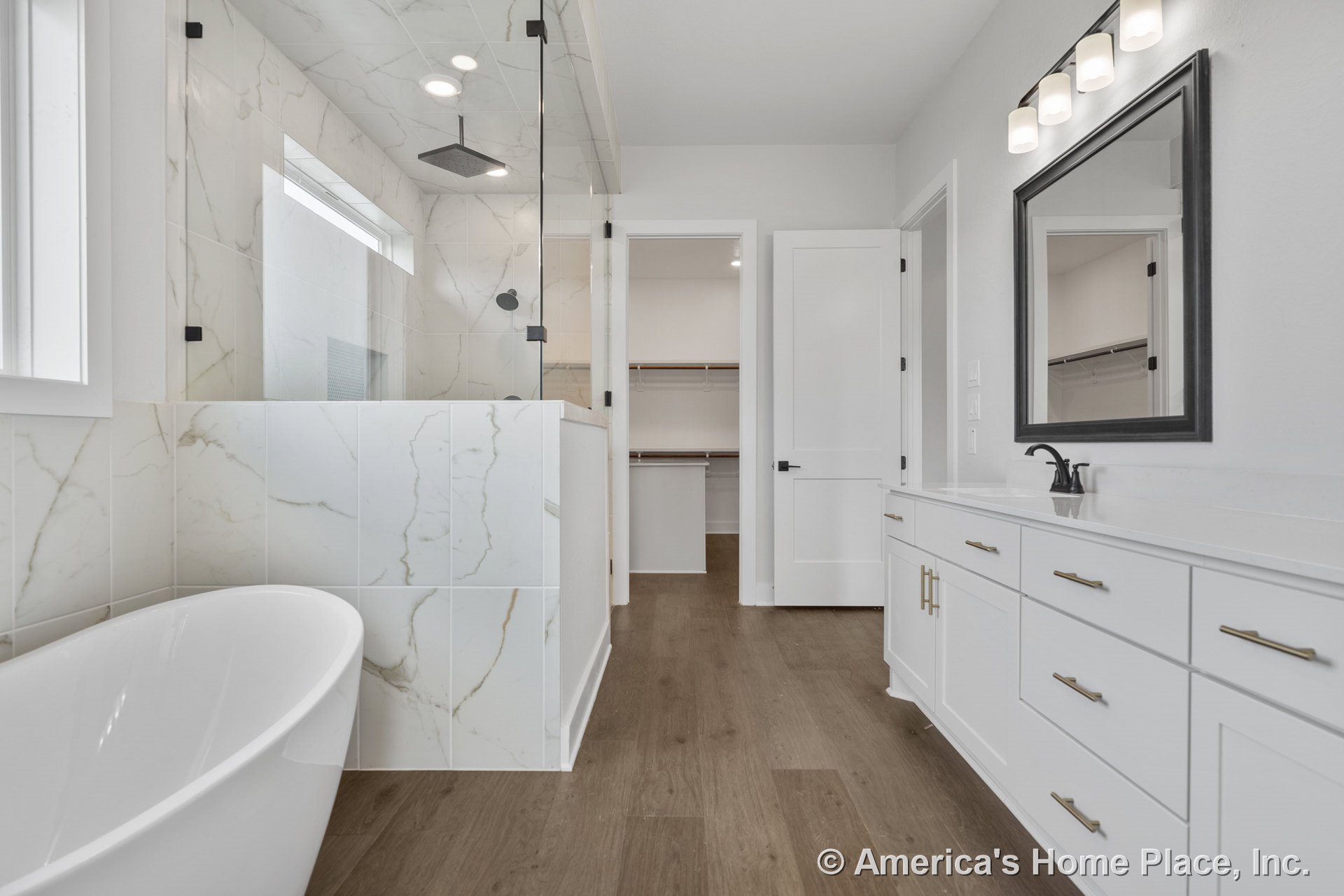 Freestanding bathtub beside marble-look tile shower, double sink vanity with built-in lighting, wood-look flooring, large framed mirror, built-in closet shelving, window, and