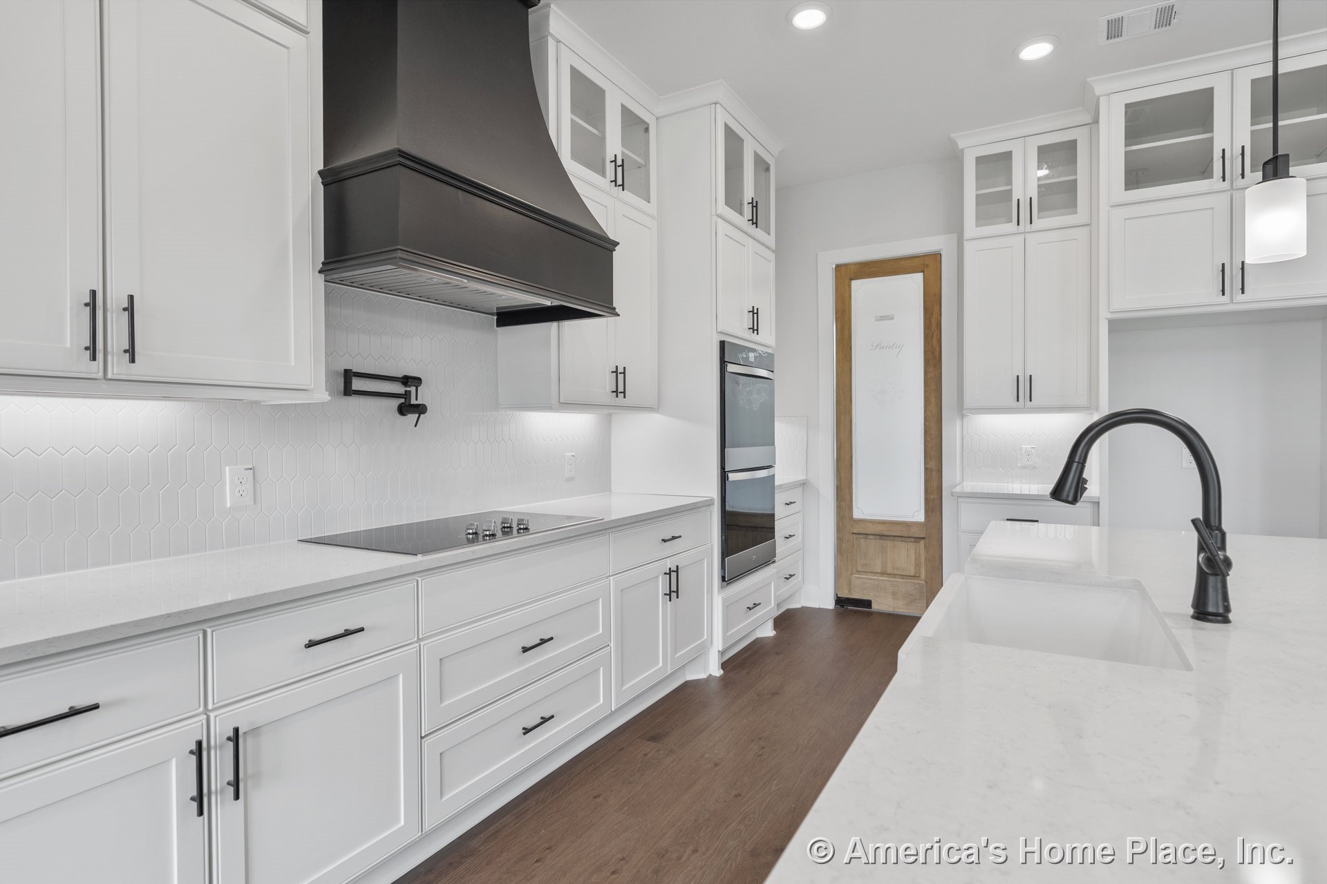 White shaker cabinets with quartz countertops, black metal range hood, hexagonal tile backsplash, built-in double oven, wood pantry door with frosted glass, undermount sink with
