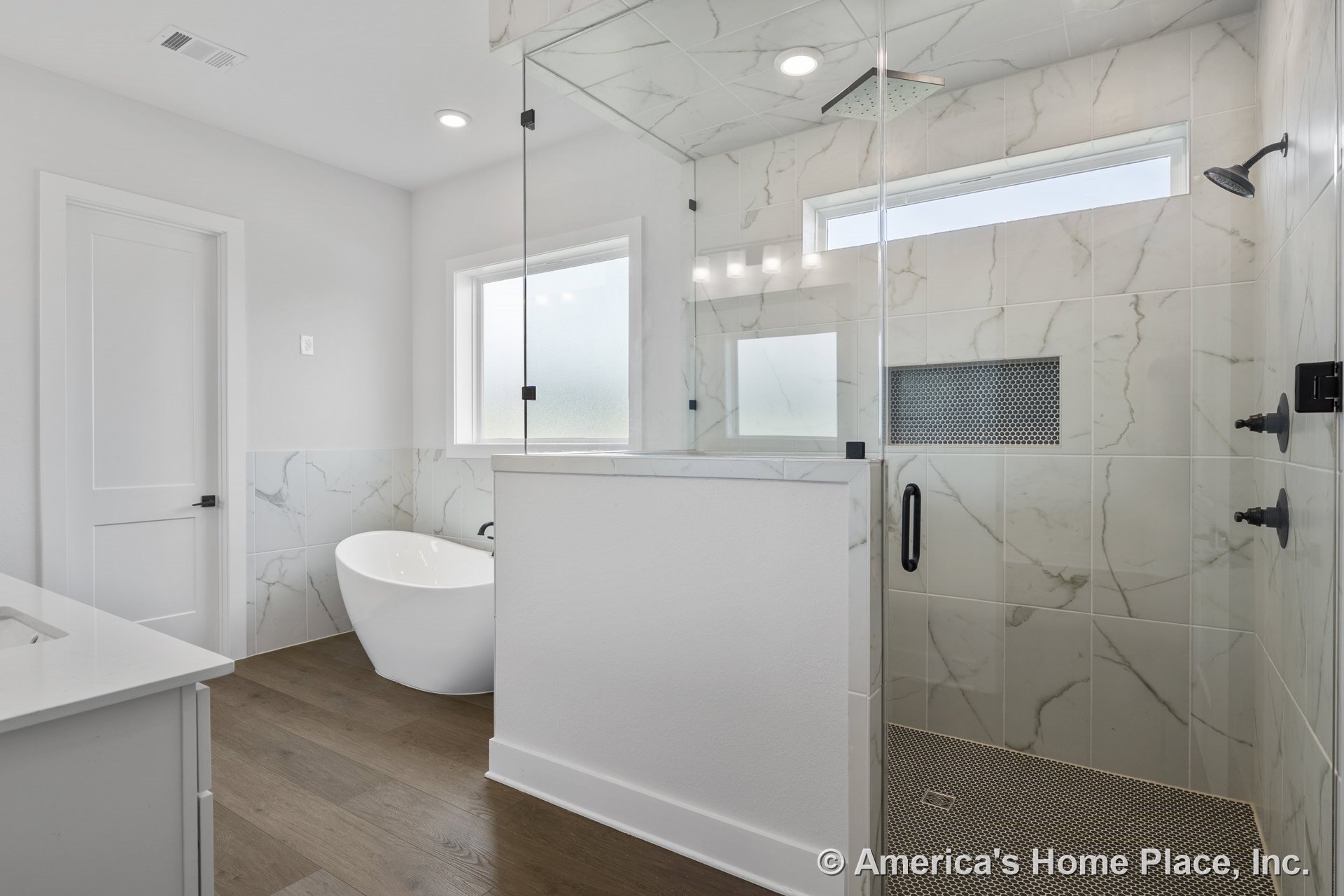 Frameless glass shower with marble-look tile walls, freestanding soaking tub, wood plank flooring, recessed ceiling lights, frosted window, shower niche, white flat-panel