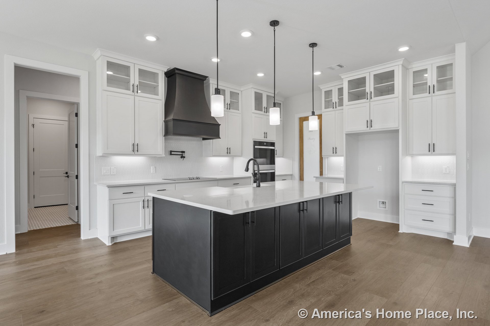 Large black kitchen island with white quartz countertop, white upper cabinets featuring glass fronts, modern pendant lights above island, built-in oven, prominent vent hood, wood