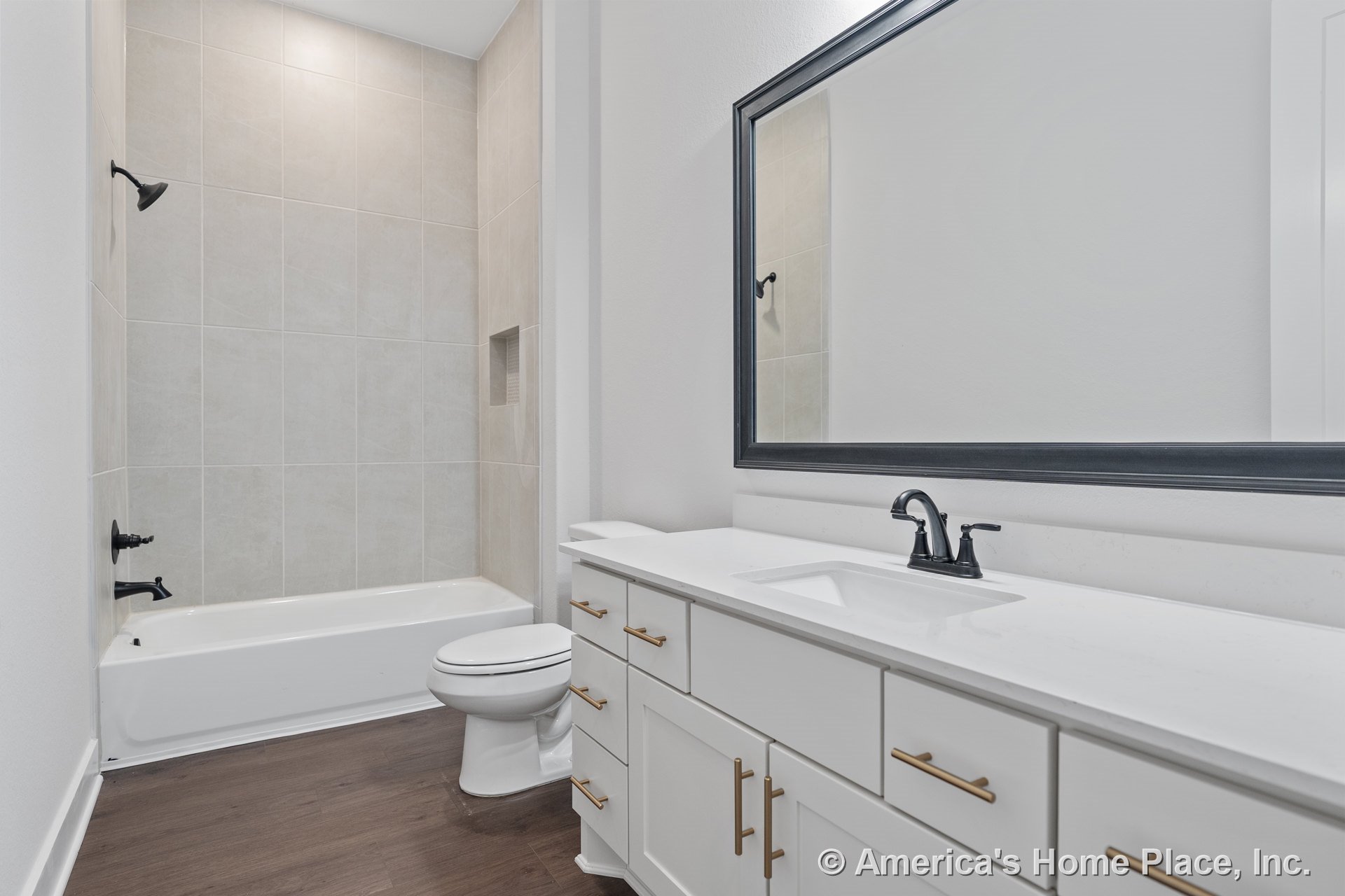 Large tiled shower wall with built-in niche, white shaker vanity topped with quartz countertop, matte black faucet, framed mirror, and wood-look flooring.