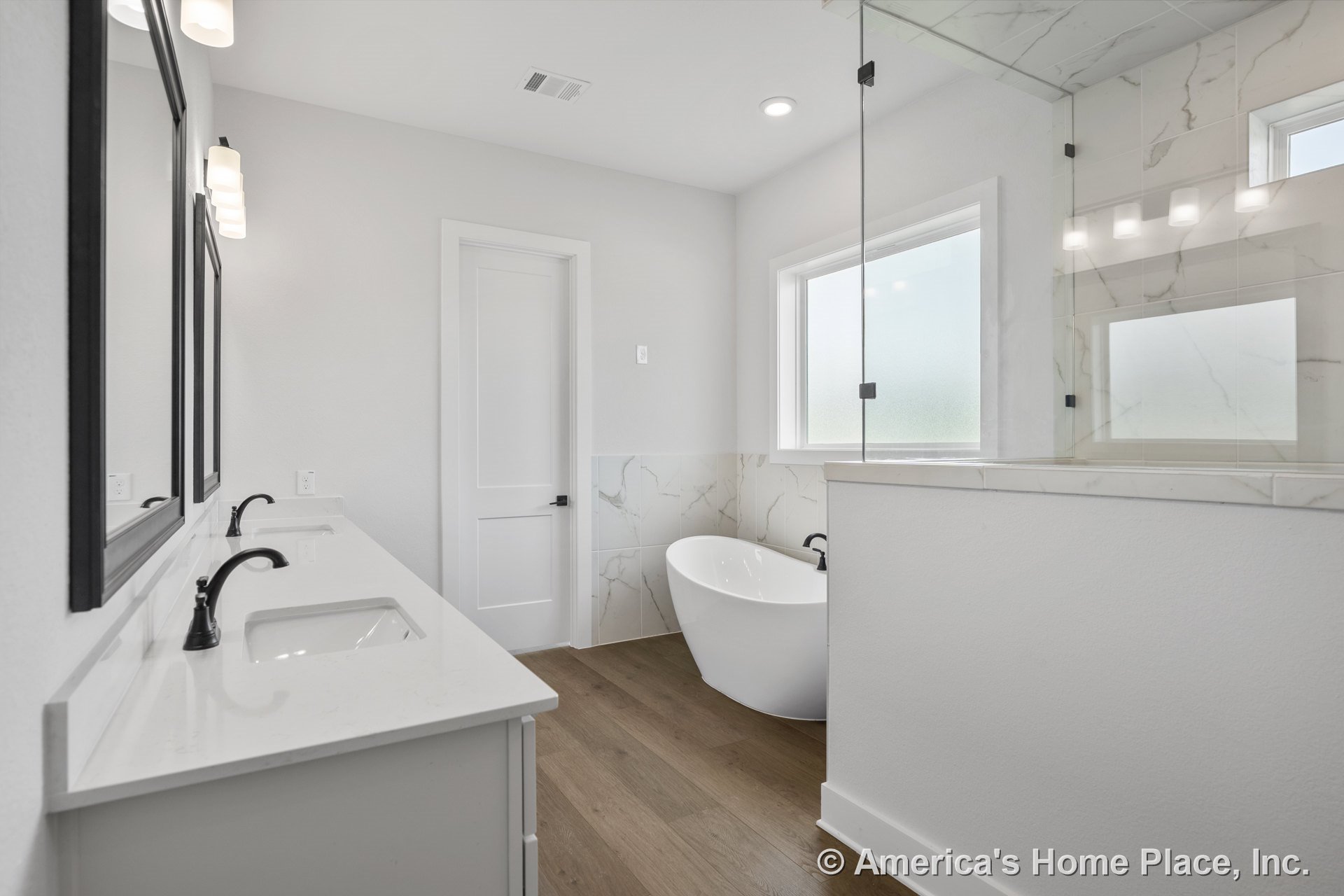 Freestanding soaking tub beside frameless glass shower with marble-look wall tile, double vanity with quartz countertop and black faucets, wood-look plank flooring, recessed