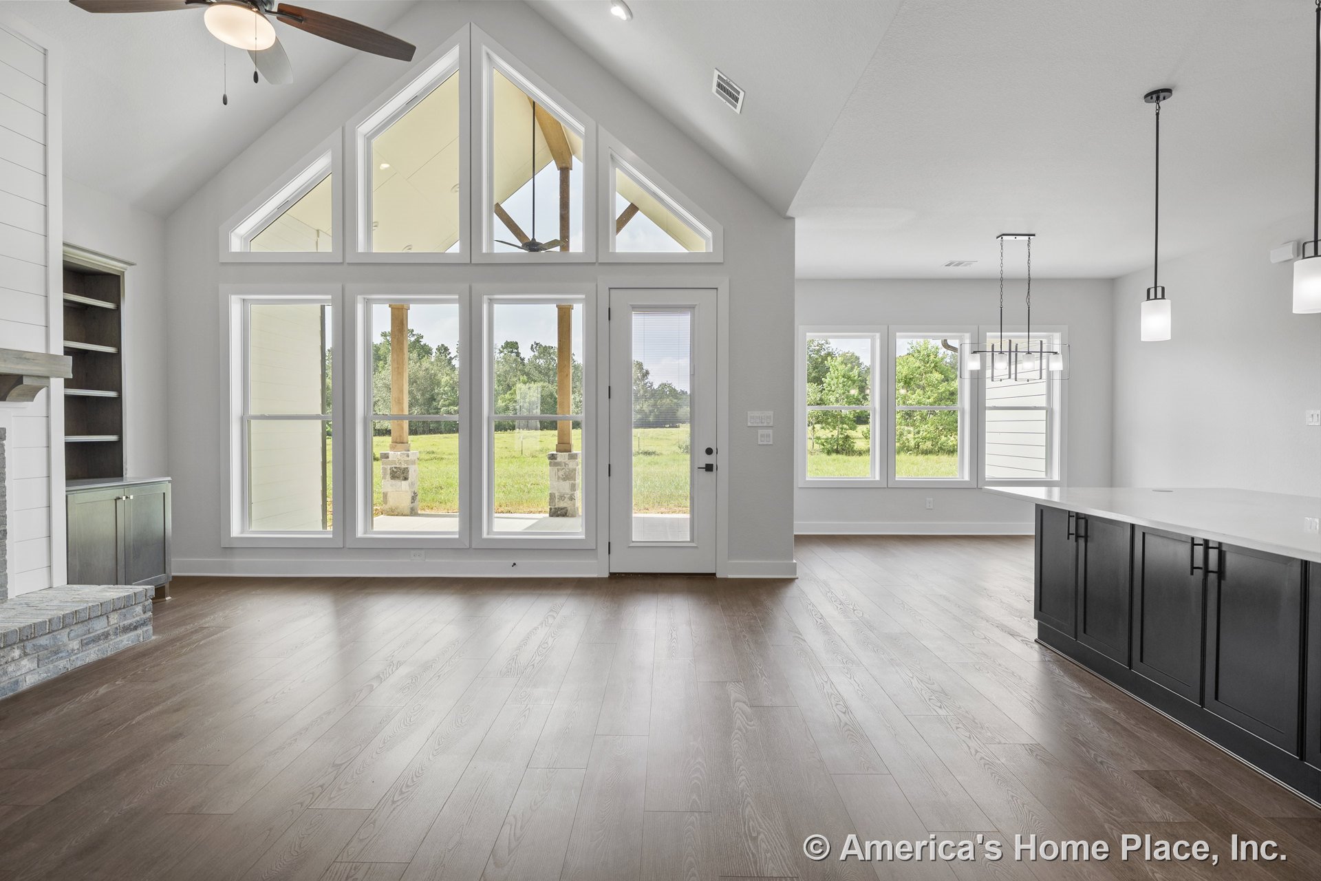 Vaulted ceiling with recessed lighting, expansive window wall and glass door, built-in shelves and cabinets flanking a stone fireplace hearth, open kitchen with pendant lights