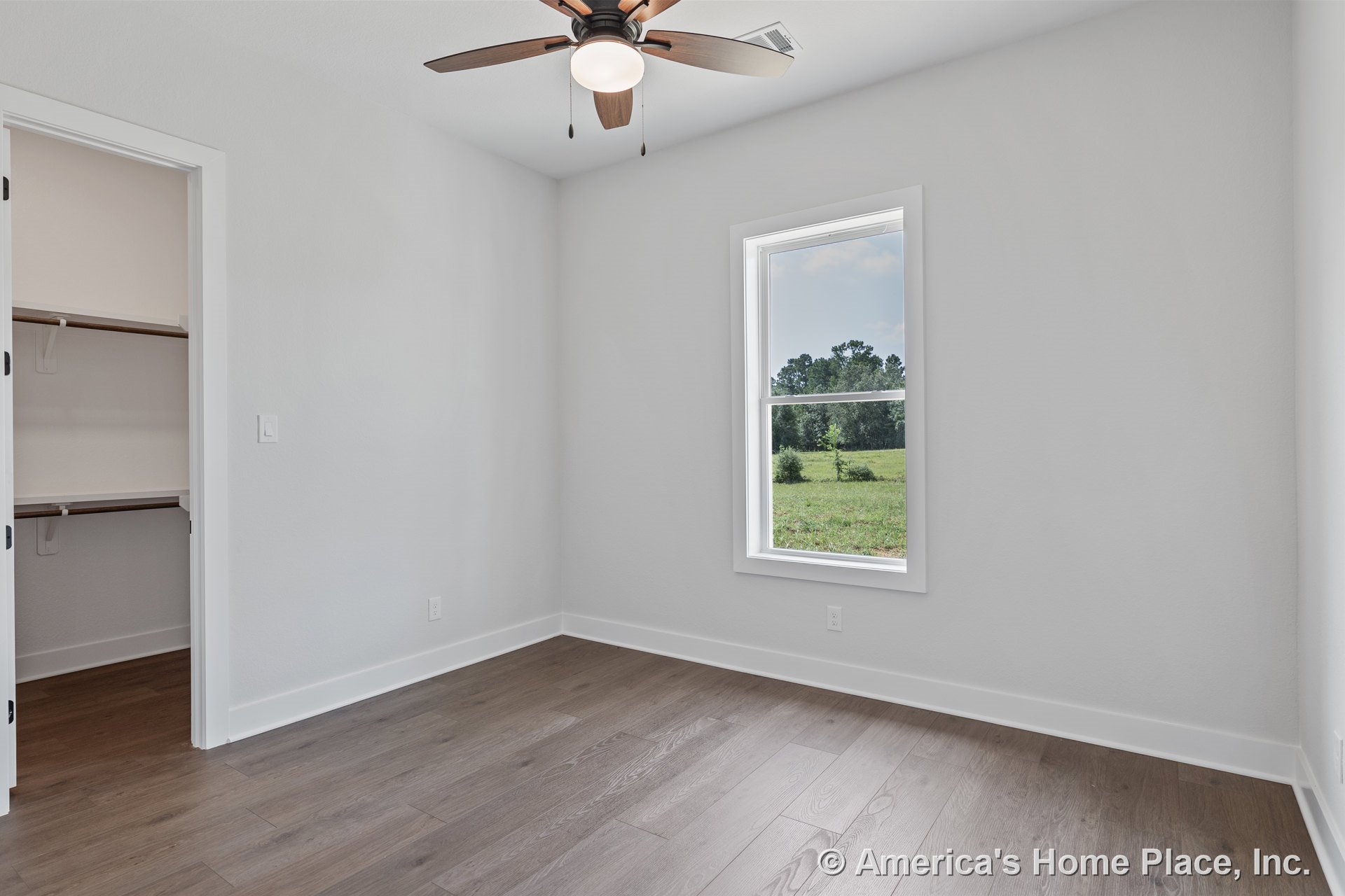 Bedroom with wood plank flooring, white painted walls, single hung window with white trim, ceiling fan and light fixture, open closet featuring built-in shelving, white baseboards