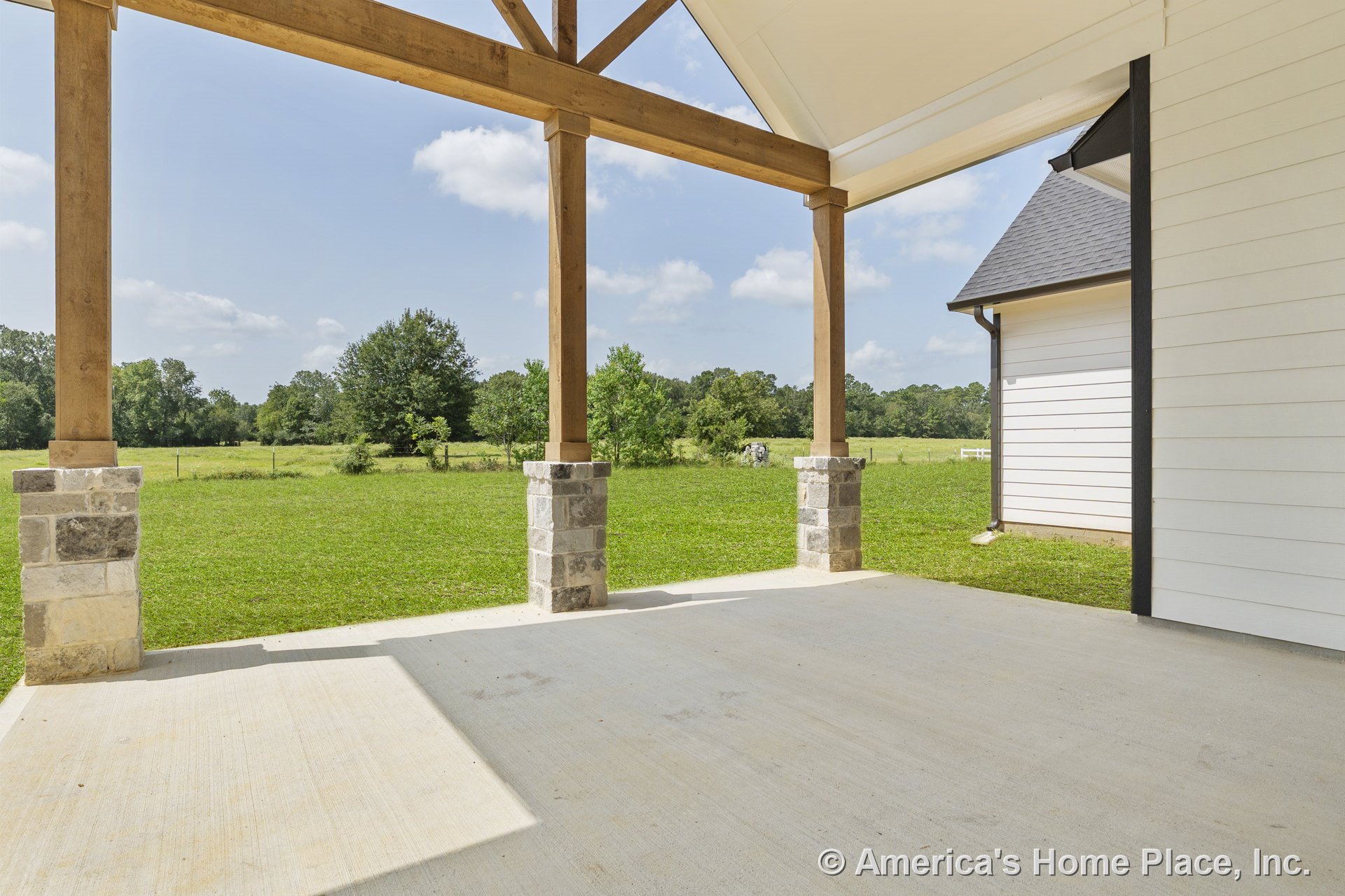 Covered patio with concrete flooring, exposed wood beams, stone column bases, open gable roof, and white siding exterior overlooking a spacious grassy yard.