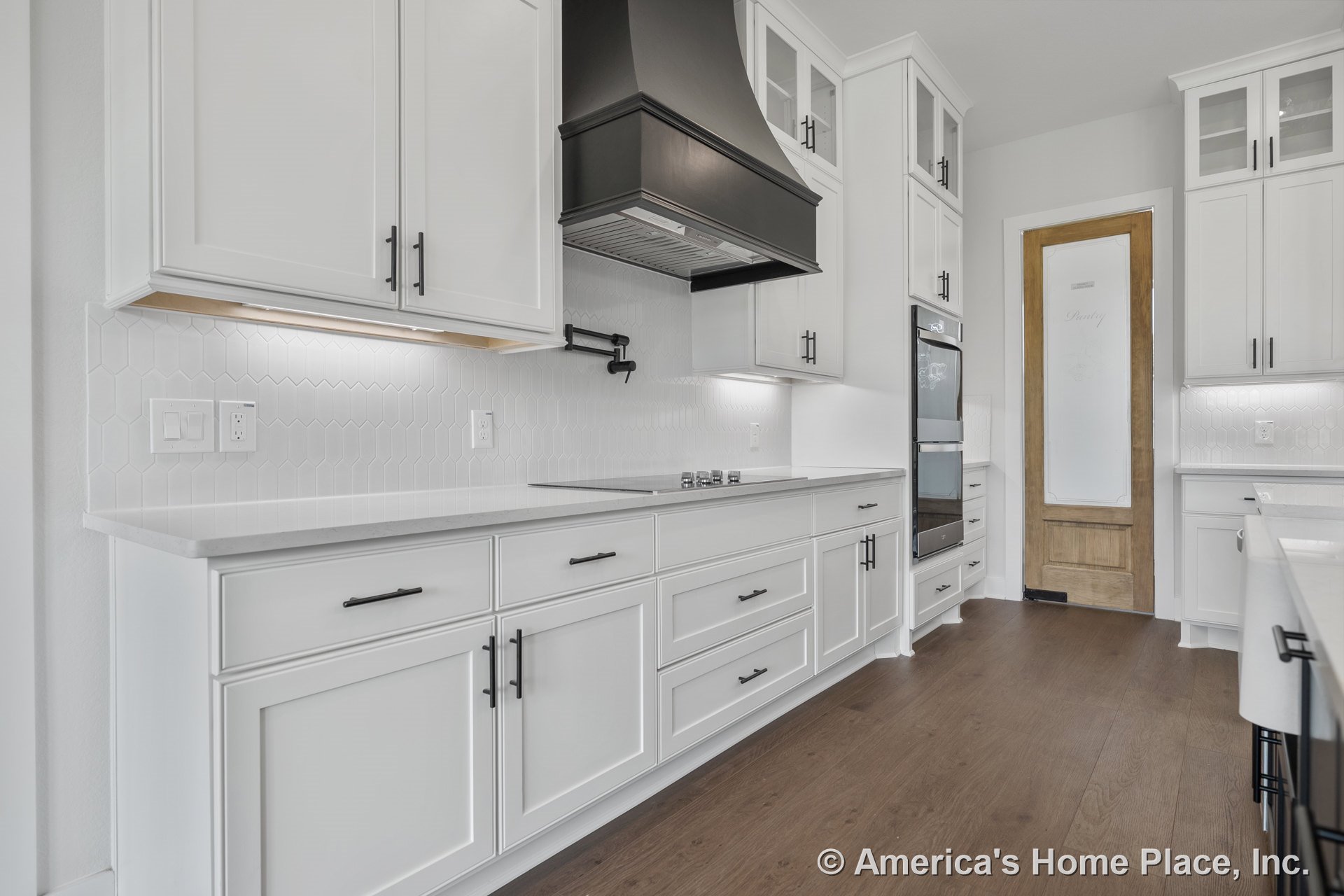 White shaker cabinets with black hardware, black metal range hood, hexagonal tile backsplash, built-in double oven, wood pantry door with frosted glass, under-cabinet lighting, and