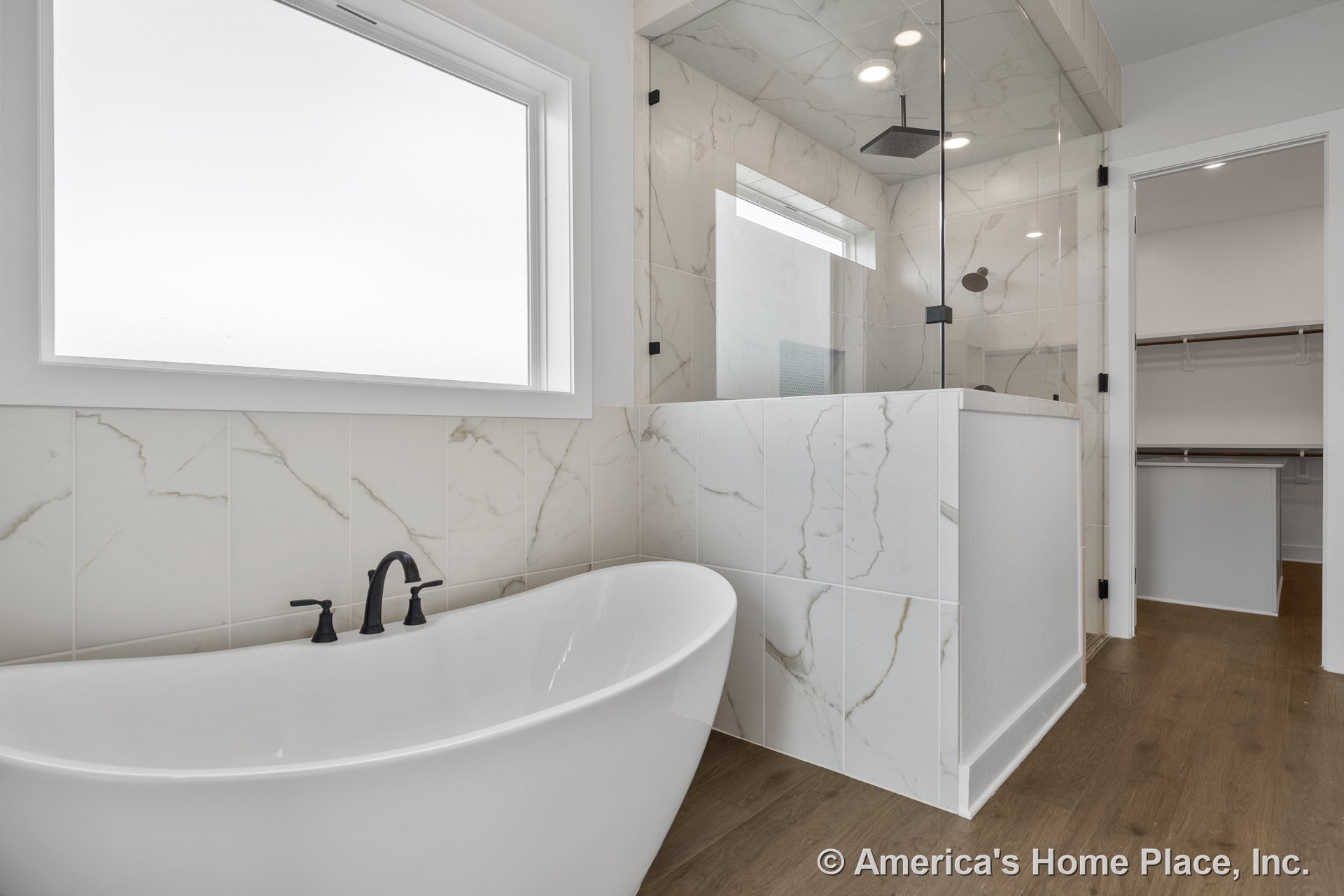 Freestanding bathtub beside marble-look tile walls, glass shower enclosure with rain showerhead and black plumbing fixtures, large frosted window, wood-look flooring, built-in