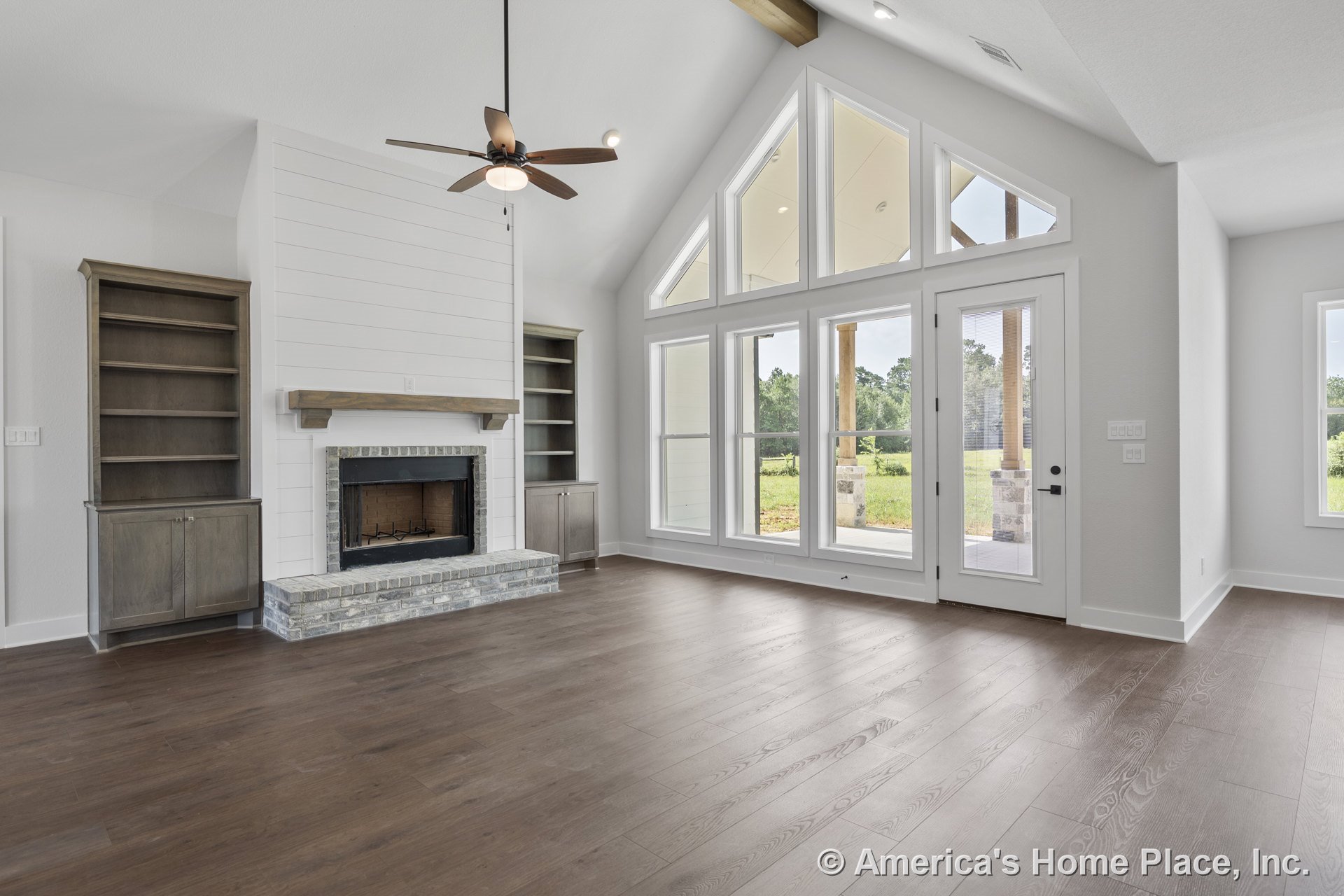 Vaulted ceiling with exposed beam, built-in wood cabinetry flanking a shiplap fireplace surround, brick hearth, wide plank wood flooring, floor-to-ceiling windows and glass door