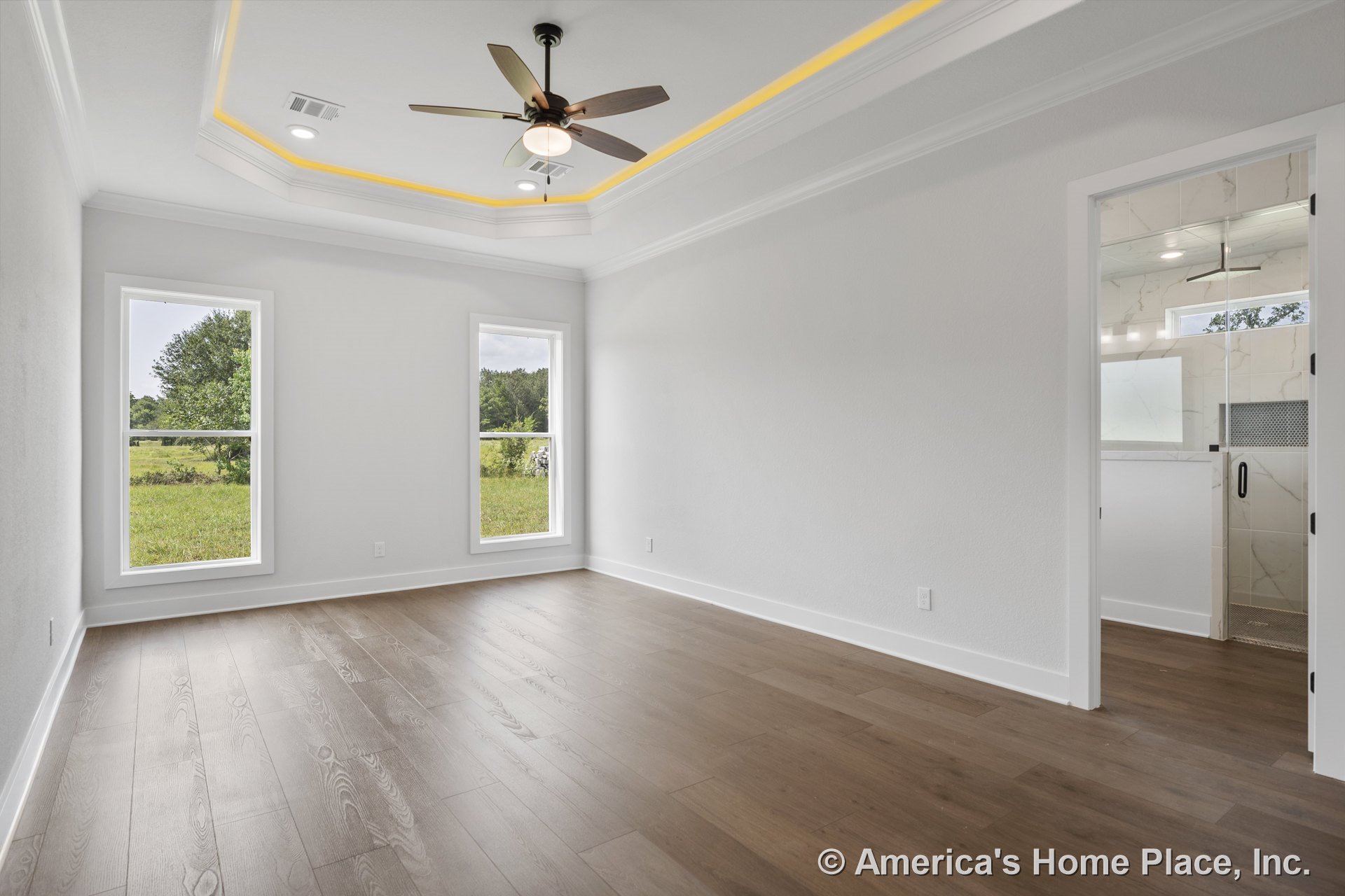 Tray ceiling with recessed LED lighting and ceiling fan, wood plank flooring, two tall windows with white trim and baseboards, bathroom entry featuring tiled walls, modern finishes