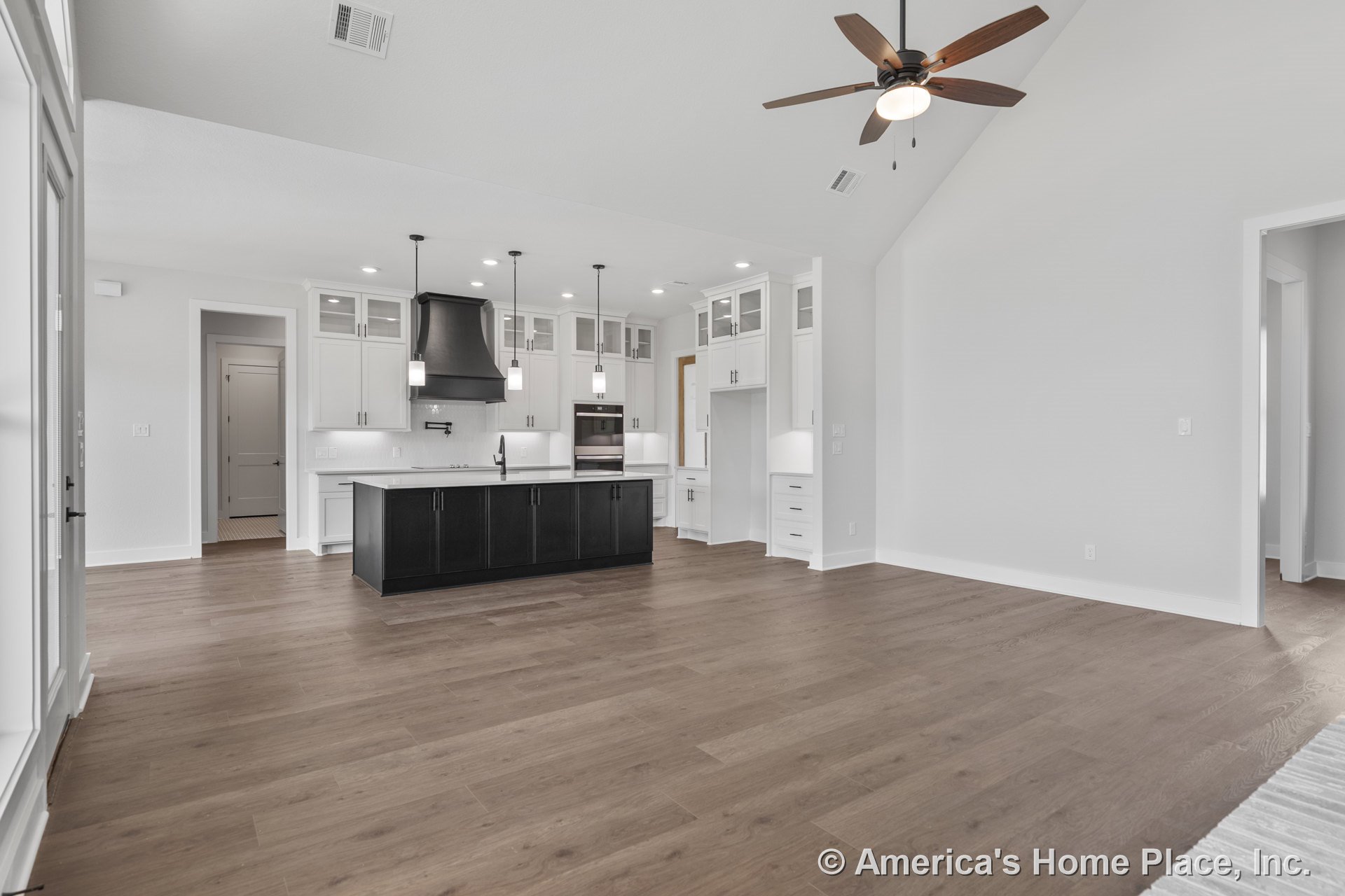 Black kitchen island with pendant lights overhead, white upper cabinets featuring glass doors, wood-look flooring, built-in oven and range hood, vaulted ceiling with ceiling fan.