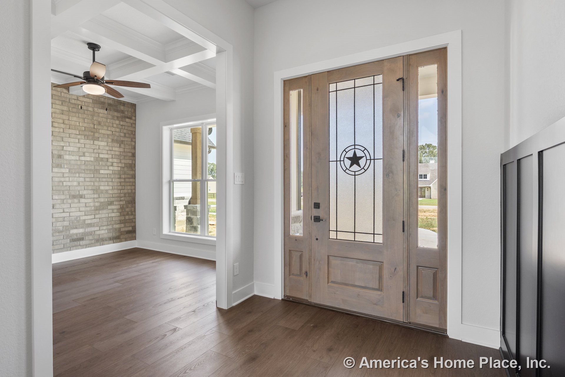 Wooden front door with decorative glass panels featuring a star motif, adjacent to a room with a brick accent wall, coffered ceiling with ceiling fan, large window with white trim