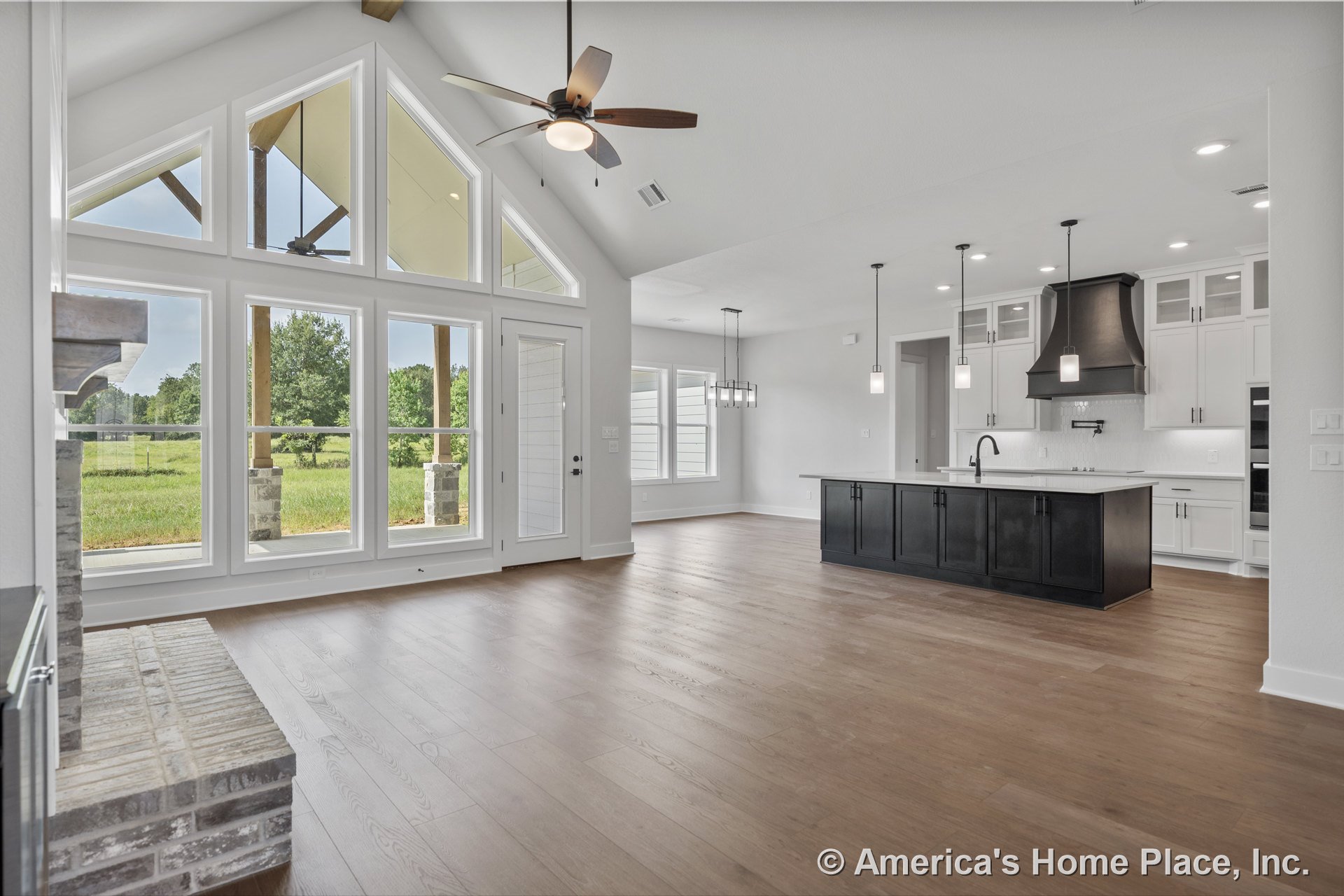 Vaulted ceiling with exposed beam, floor-to-ceiling picture windows, brick fireplace hearth, wide plank wood flooring, modern kitchen featuring black island with pendant lights and