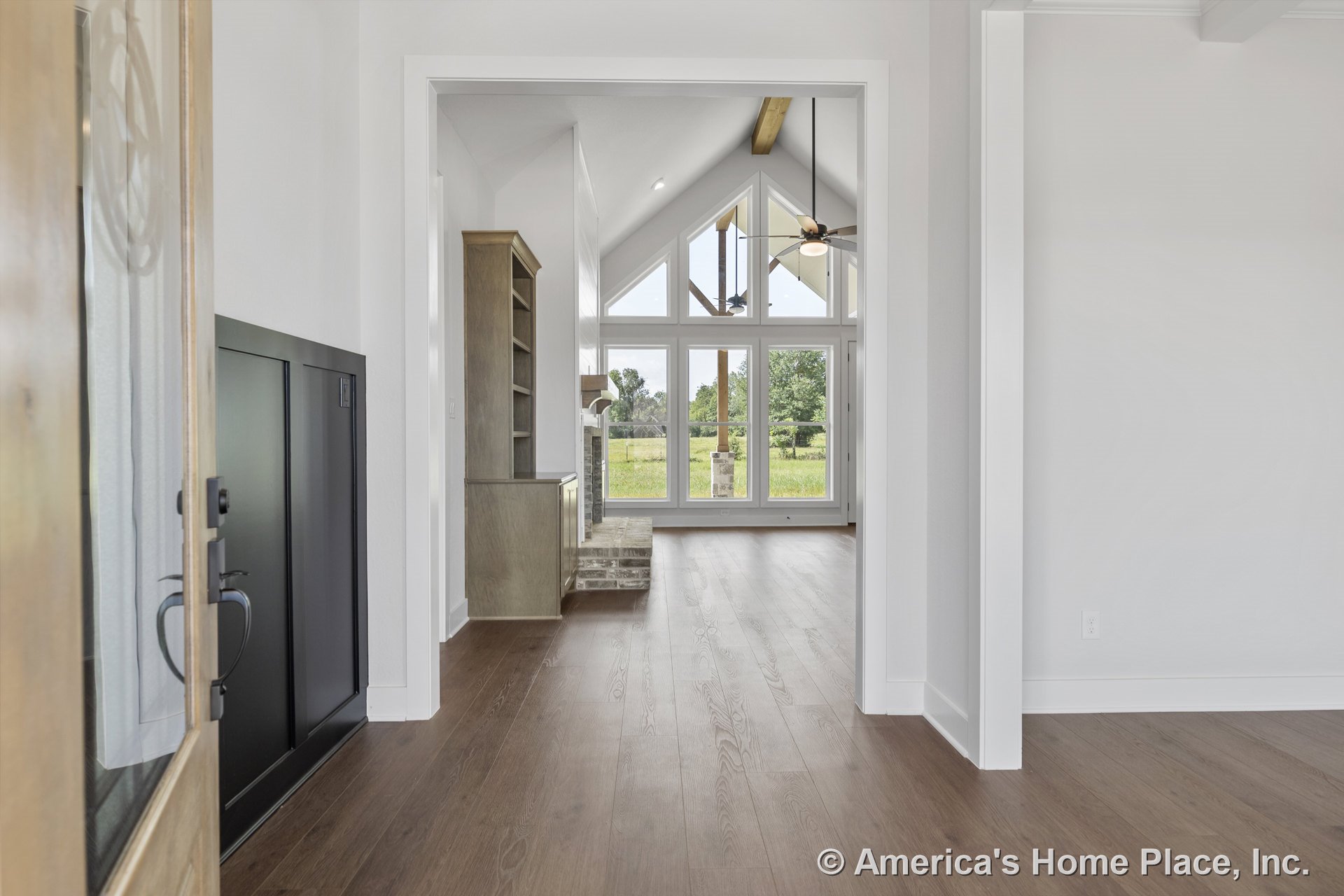 Vaulted living room ceiling with exposed wood beams, expansive window wall, built-in shelving flanking a fireplace surround, wood flooring, and detailed trim; foyer entry visible