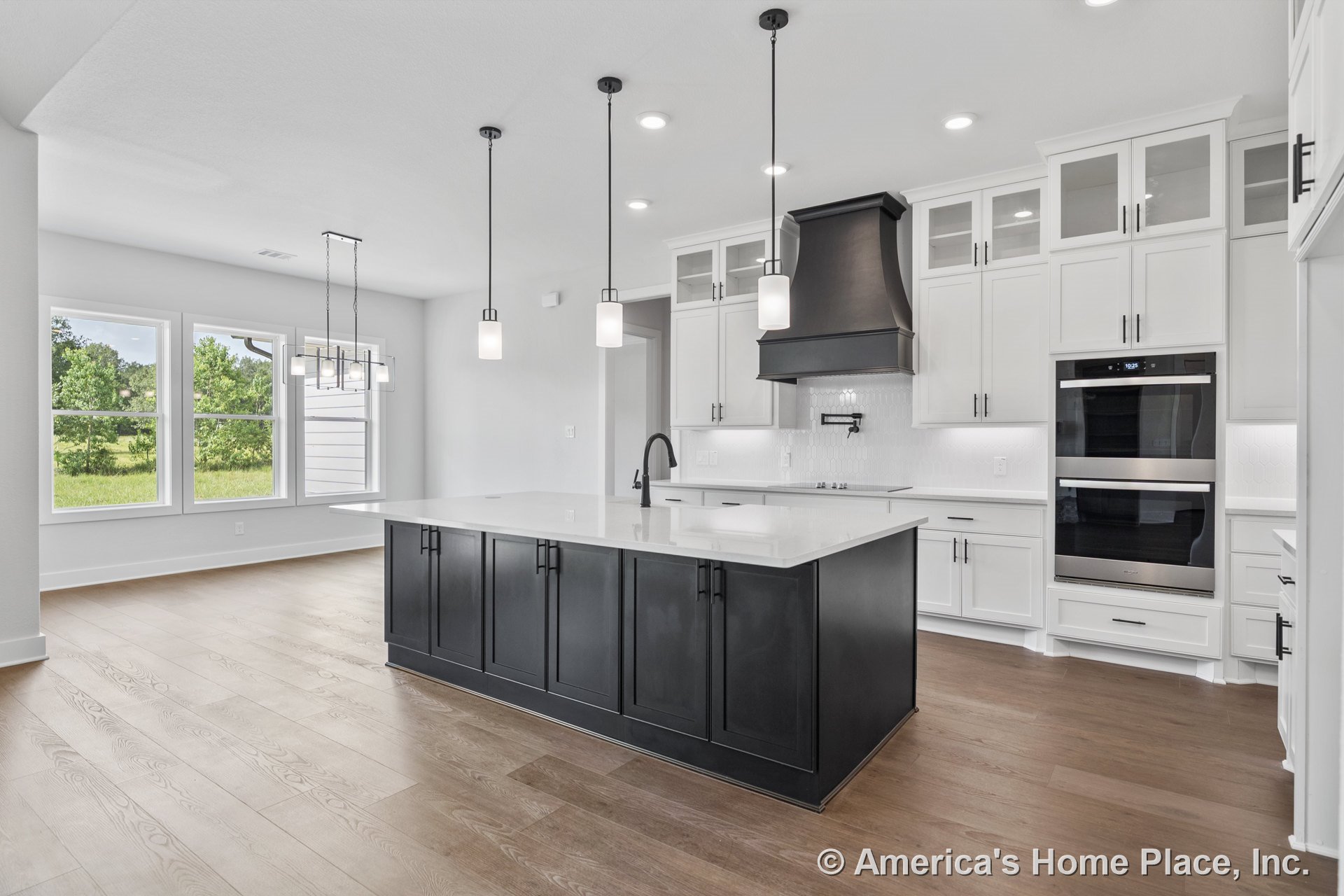 Spacious kitchen with black island cabinetry topped by white quartz countertops, white upper and lower cabinets, double wall ovens, pendant lighting fixtures, wood plank flooring