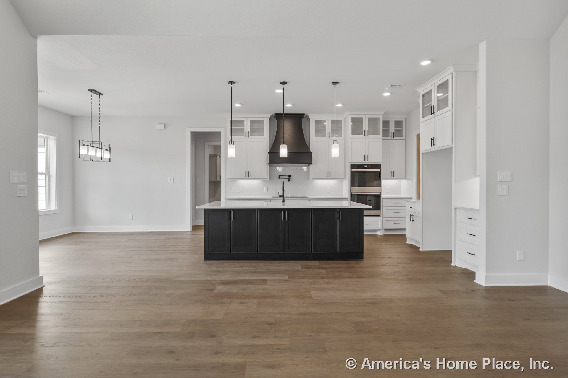 Spacious open kitchen with large central island featuring dark cabinetry, white shaker upper cabinets, wood plank flooring, black range hood, built-in oven, modern pendant lights