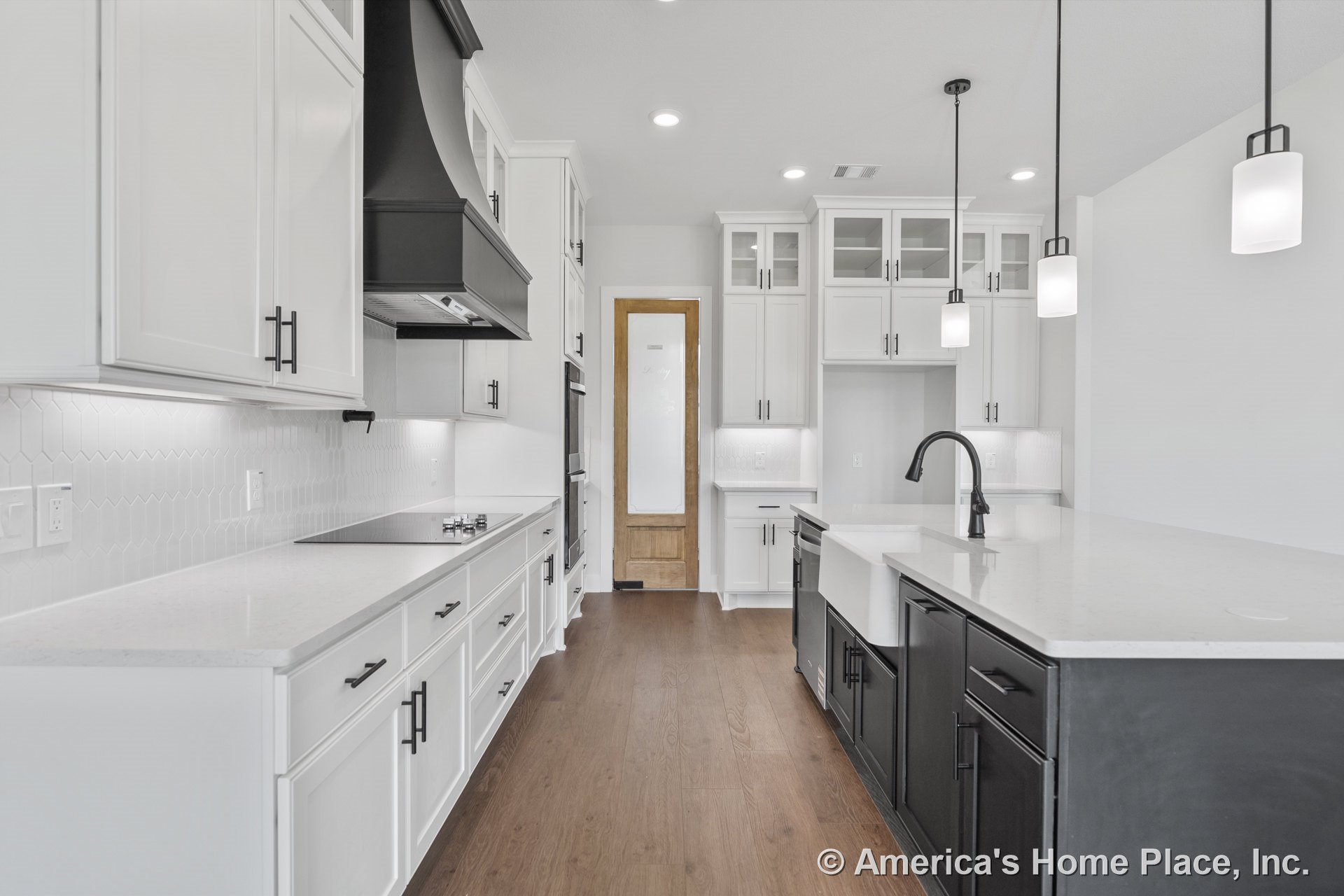 White shaker cabinets with glass doors, black range hood, quartz countertops, hexagonal tile backsplash, farmhouse sink, wood flooring, pendant lights, built-in appliances, and a