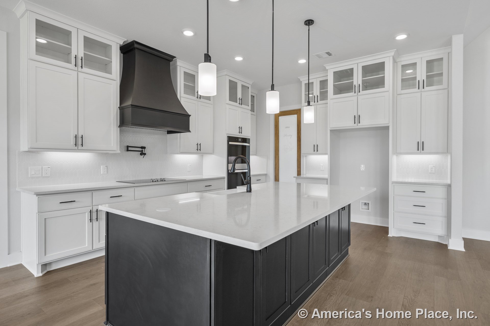 White kitchen with glass-front upper cabinets, large quartz island, black range hood, hexagonal tile backsplash, built-in oven, pot filler above cooktop, black pendant lights